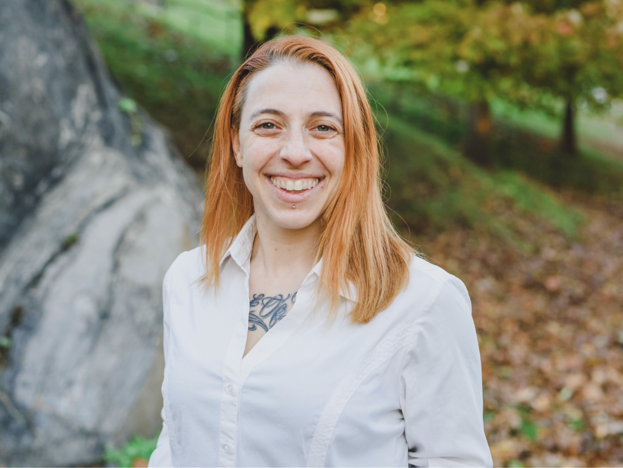 A smiling woman with red hair and a white shirt standing outdoors in a wooded area.
