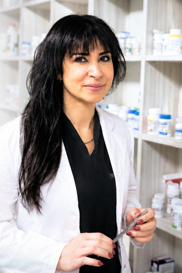 A female pharmacist standing in a pharmacy, holding a tablet, with shelves of medicine bottles in the background.