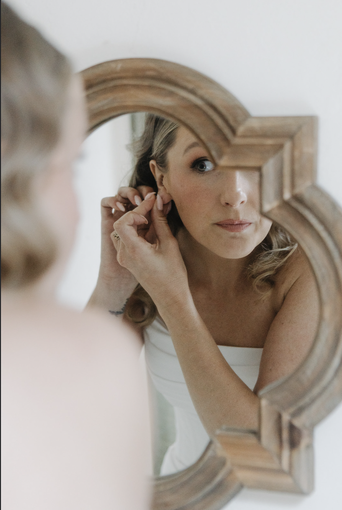 A bride getting ready for her wedding while looking in a wooden mirror, putting her earrings on, shot by two wedding photographers.