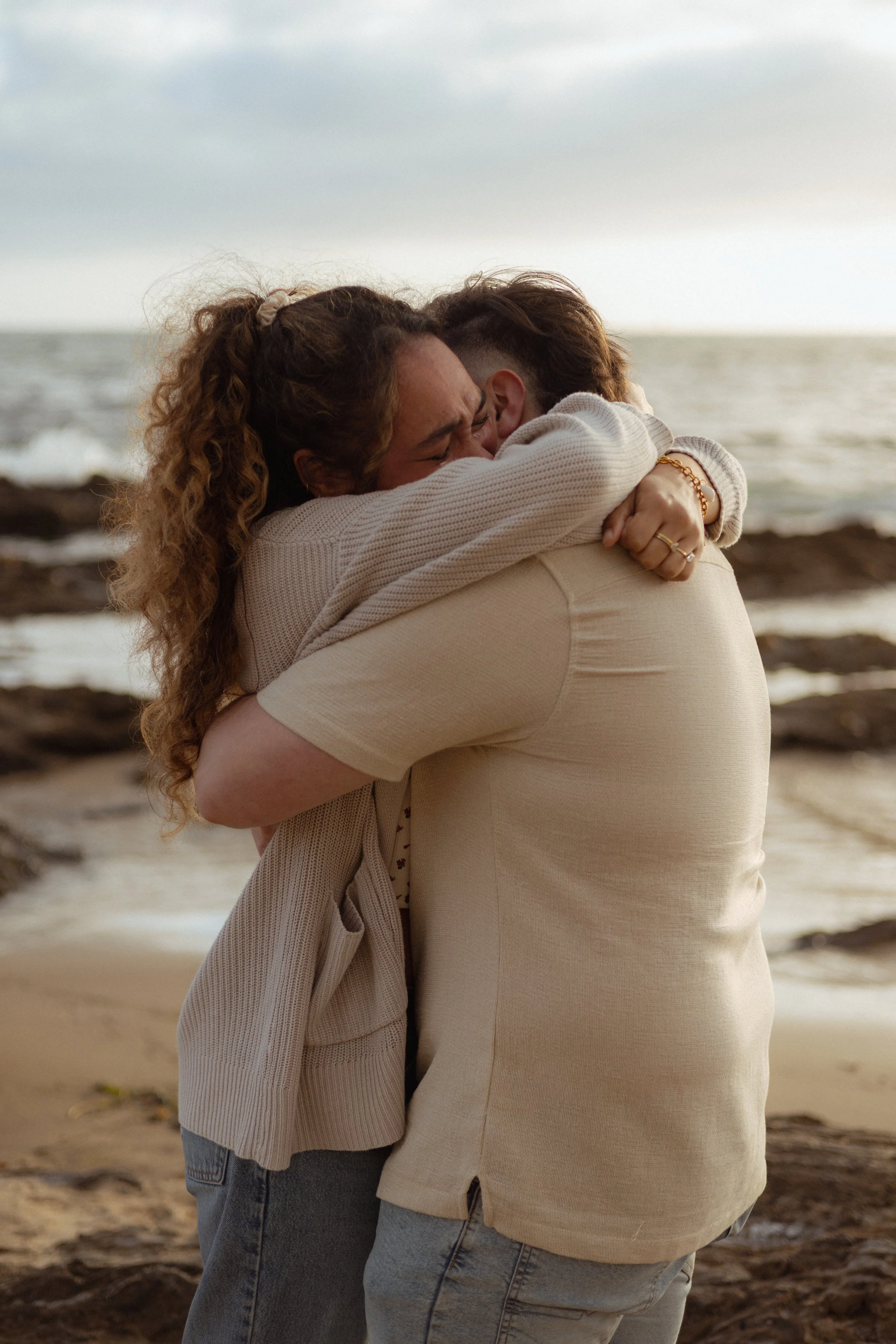 Two women hugging on a beach with ocean waves in the background, one with curly hair and the other with short hair, both smiling and appearing happy.