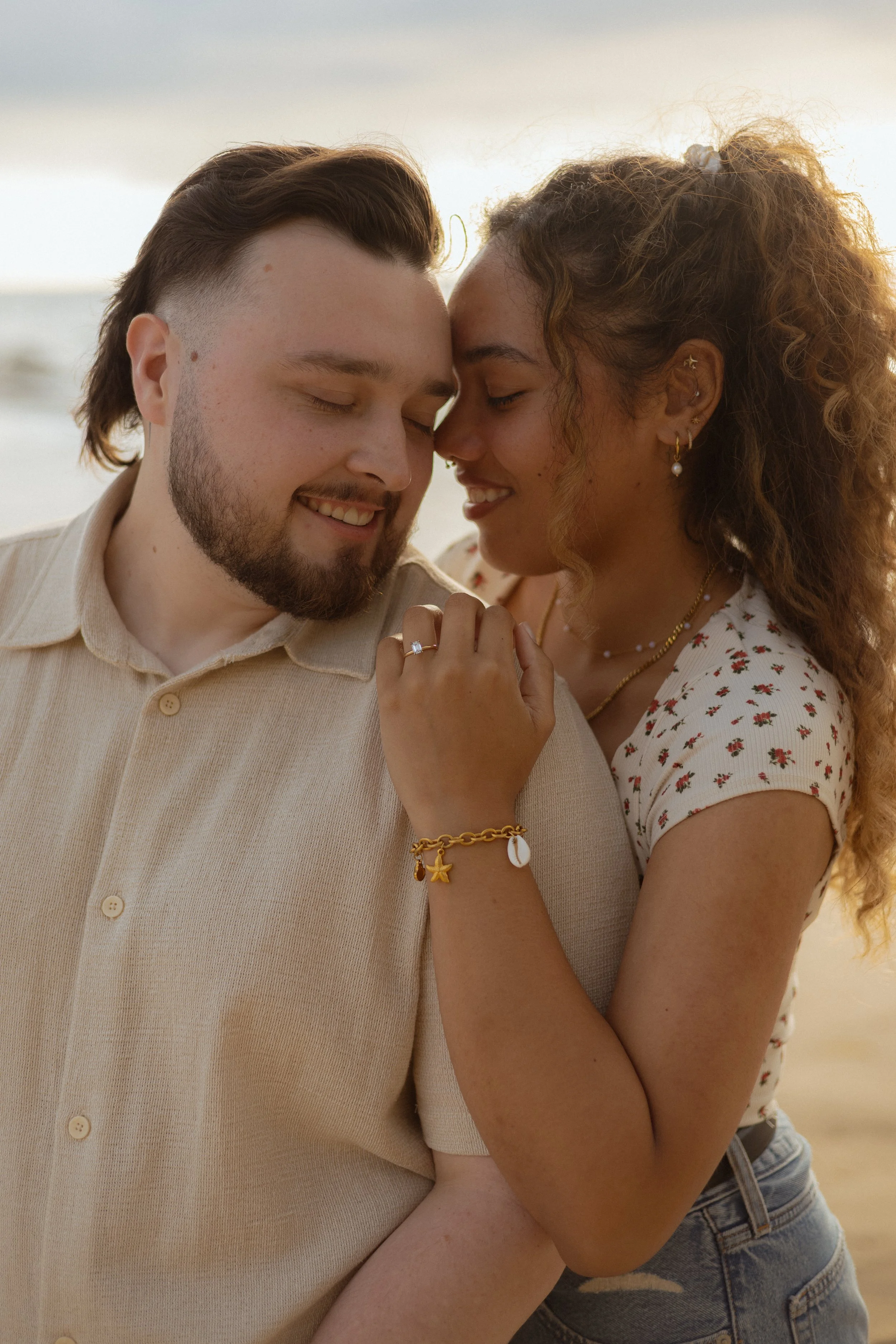 A happy couple close together with their foreheads touching on a beach at sunset, smiling with their eyes closed.