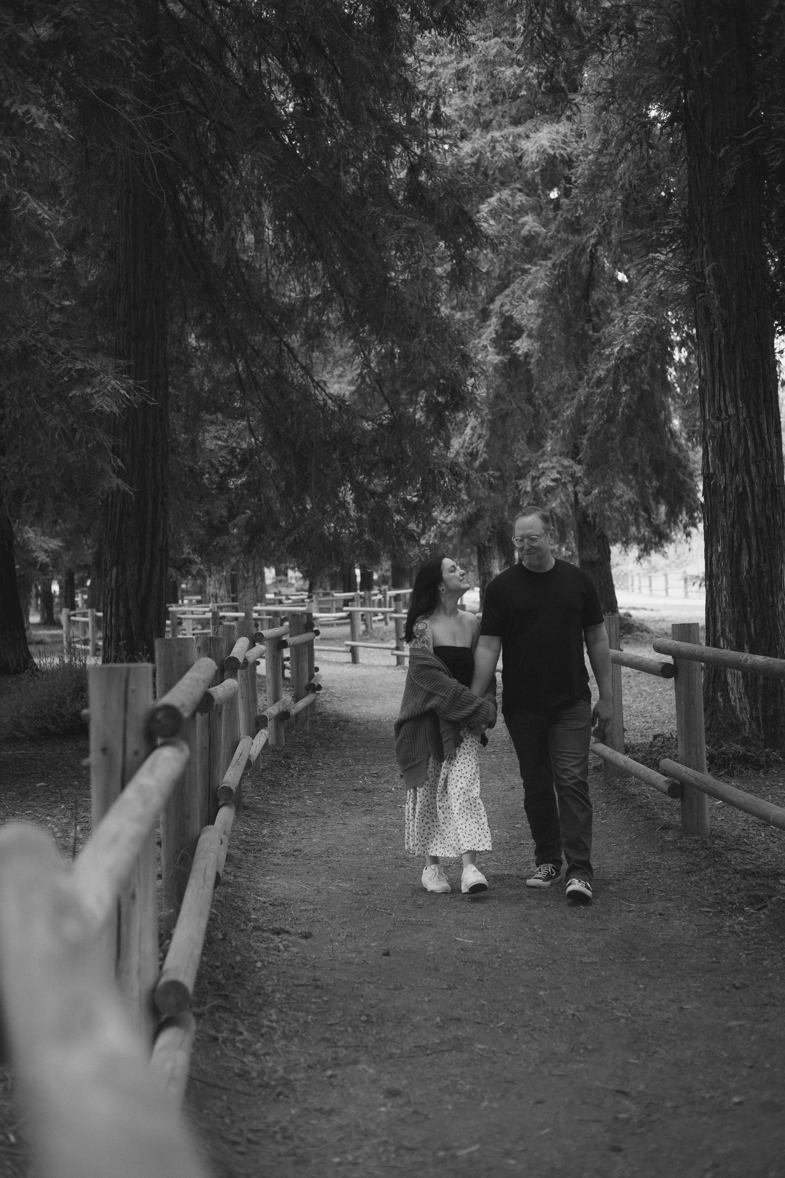 A black and white photo of a couple walking on a dirt path in a park surrounded by tall trees. The woman is looking up at the man, who is looking down at her. The scene is peaceful and intimate.