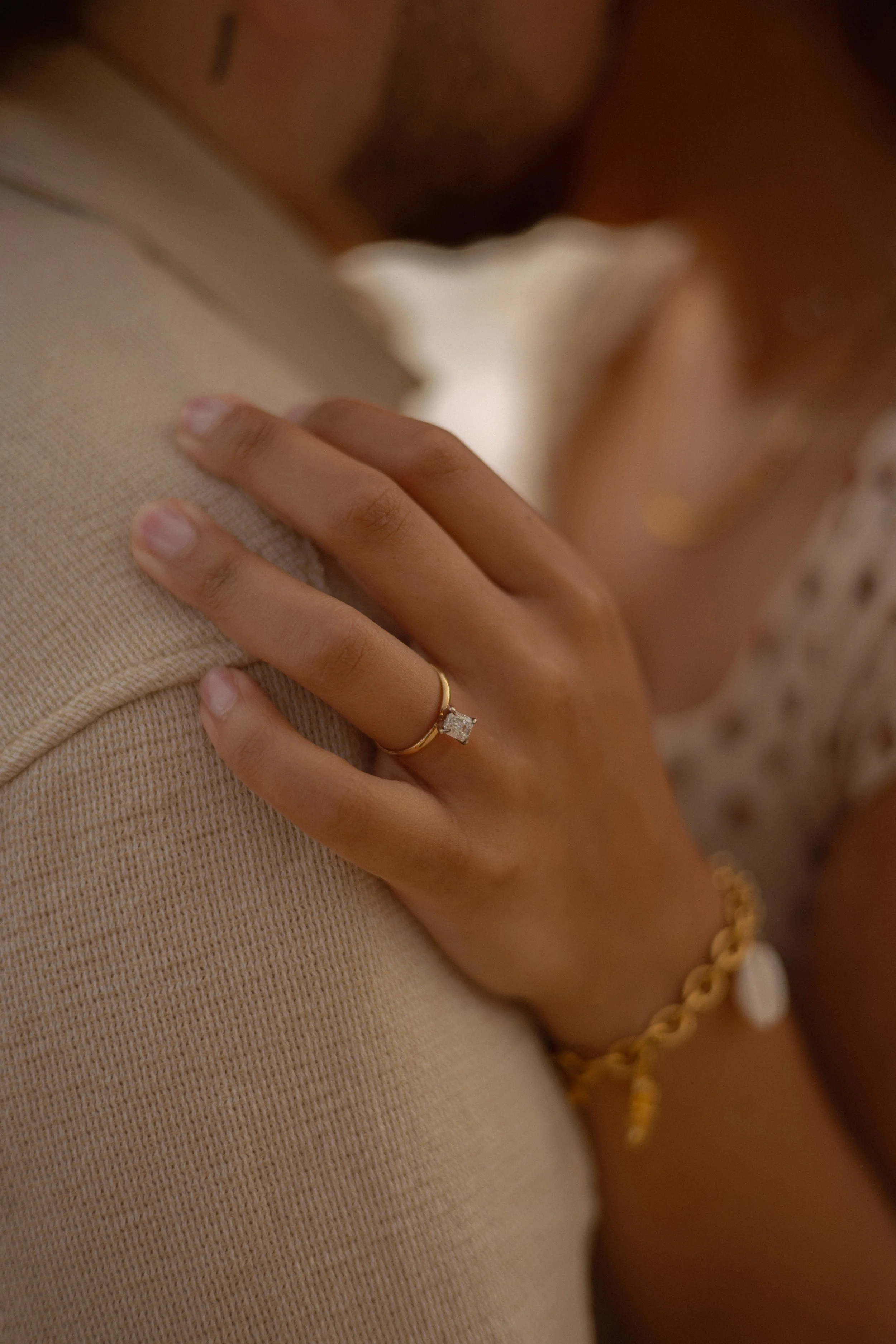 Close-up of a woman embracing a man, with her hand on his chest, showing an engagement ring, wearing a gold bracelet and jewelry.