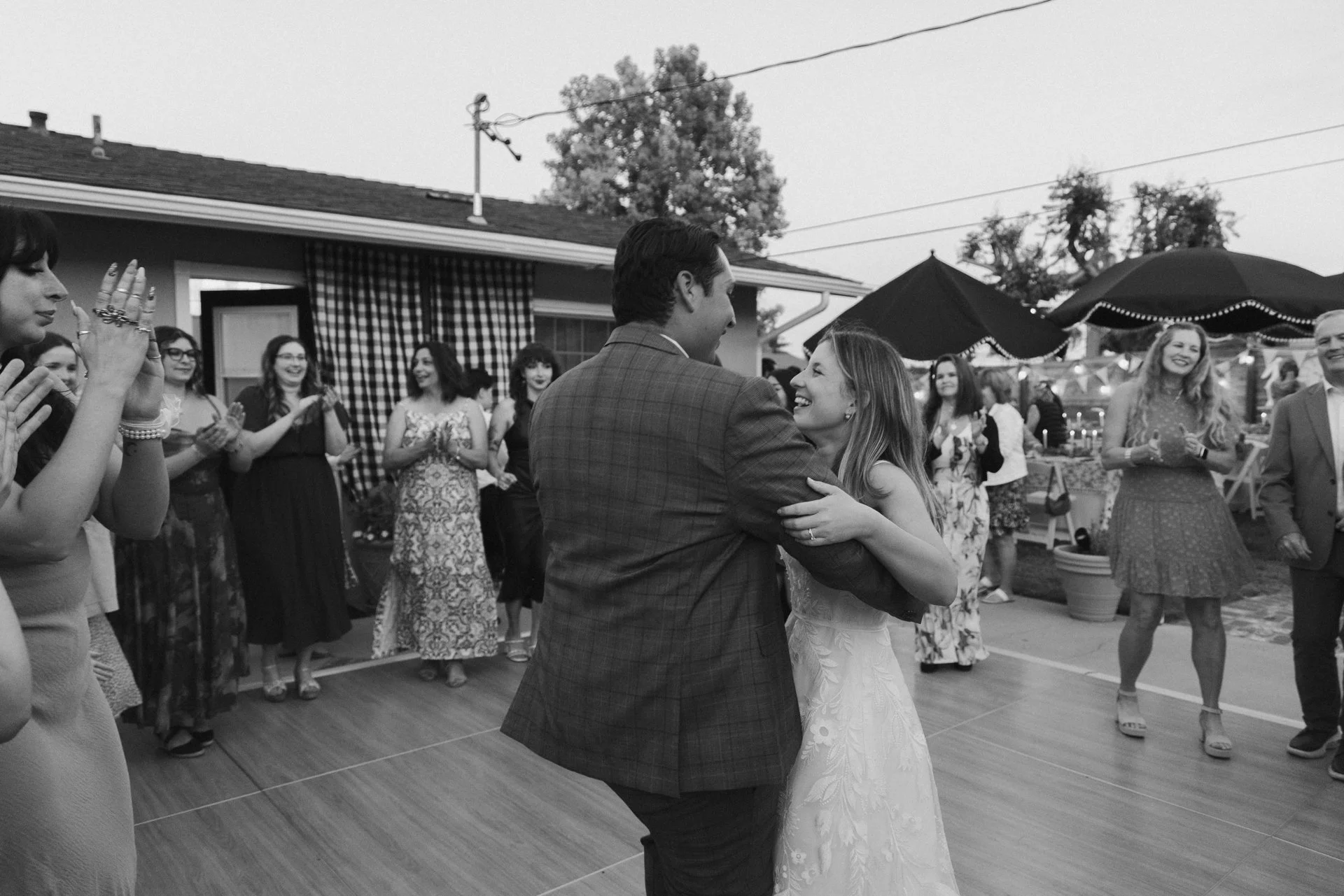 A bride and groom are dancing together during their wedding reception, surrounded by guests who are watching and smiling outdoors.