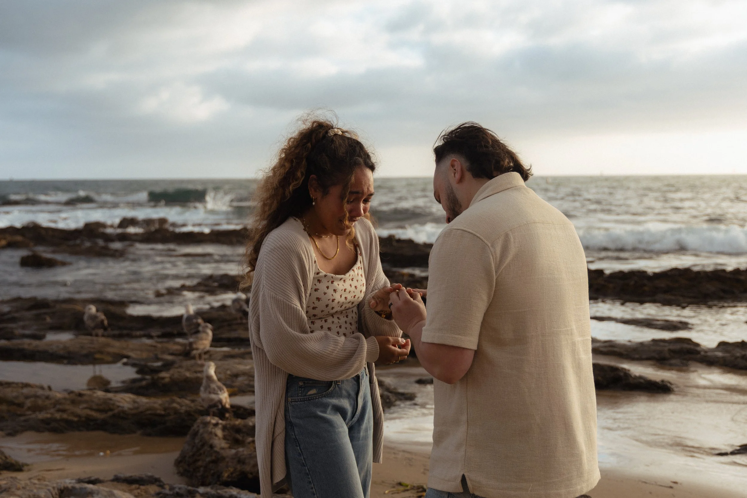 A couple is standing on a rocky beach during sunset, holding hands and looking at each other emotionally.