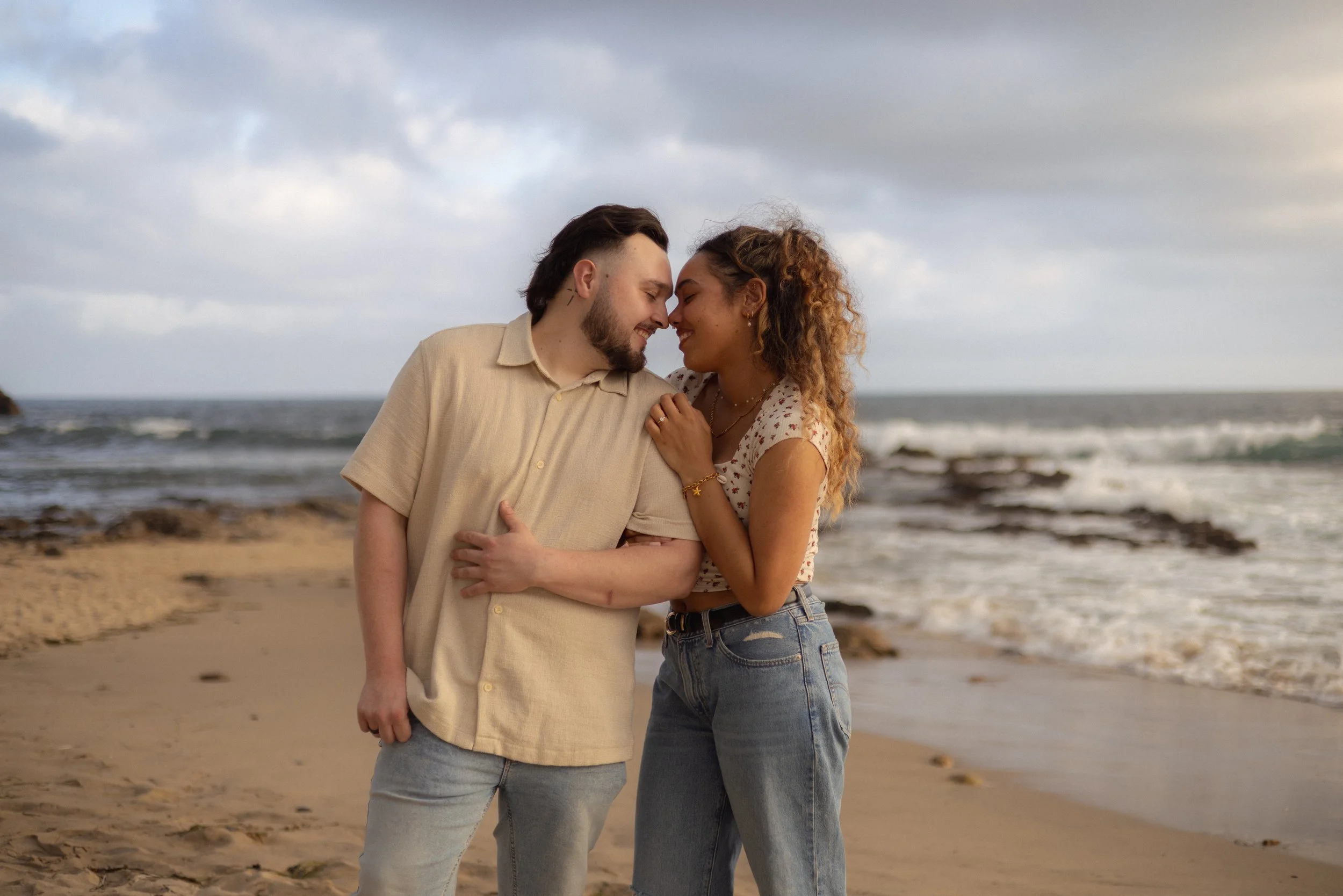 A couple standing close together on a beach, touching foreheads with eyes closed, smiling. The man has dark hair and a beard, wearing a beige shirt and jeans. The woman has curly hair, wearing a white floral top and jeans. The ocean and cloudy sky ar