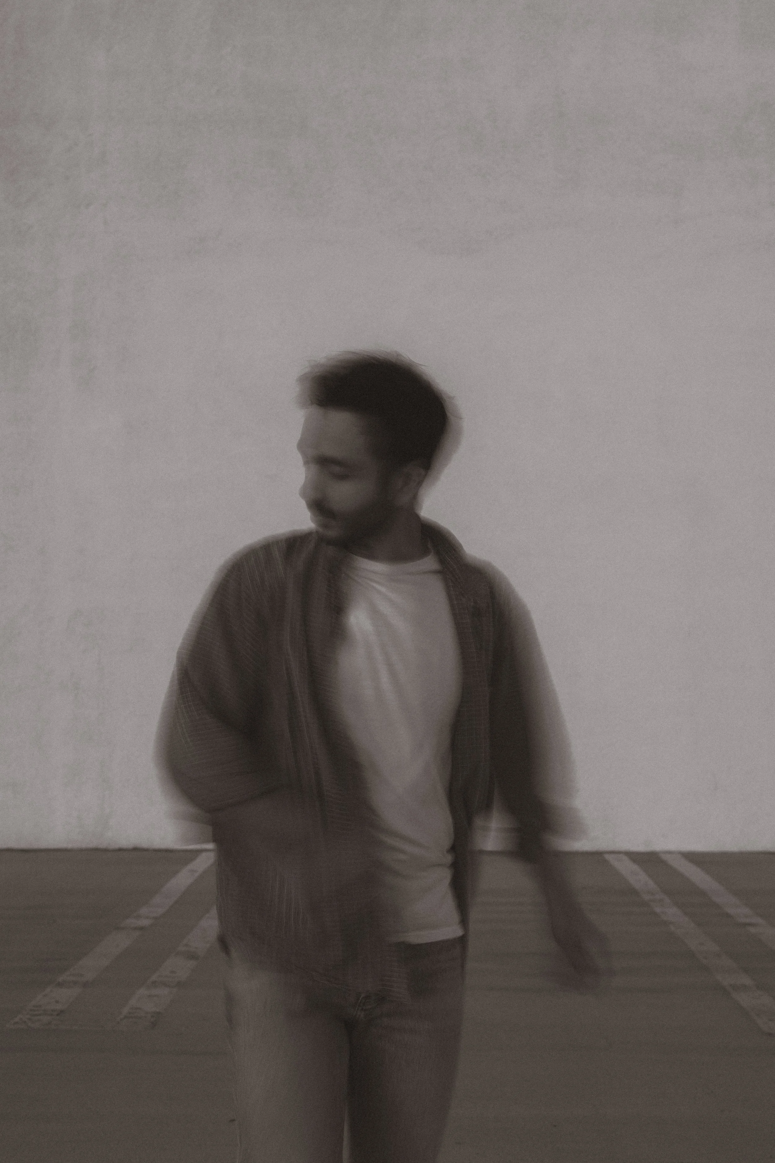 A young man with dark hair and a beard is standing in front of a plain wall and looking down. He is wearing a striped jacket over a white t-shirt.