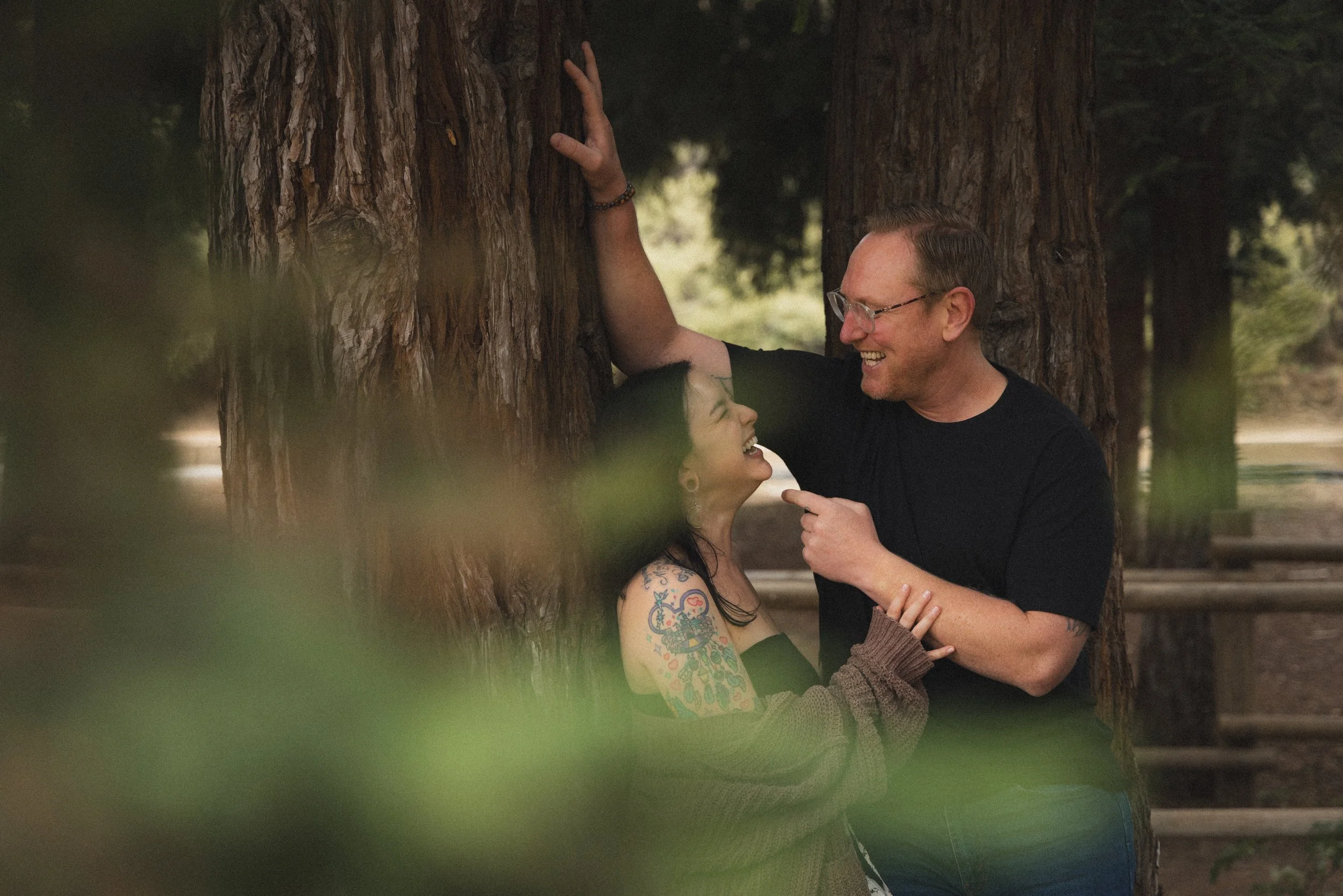 A man with glasses and a woman with tattoos smiling and hugging in a forest, with large trees in the background.
