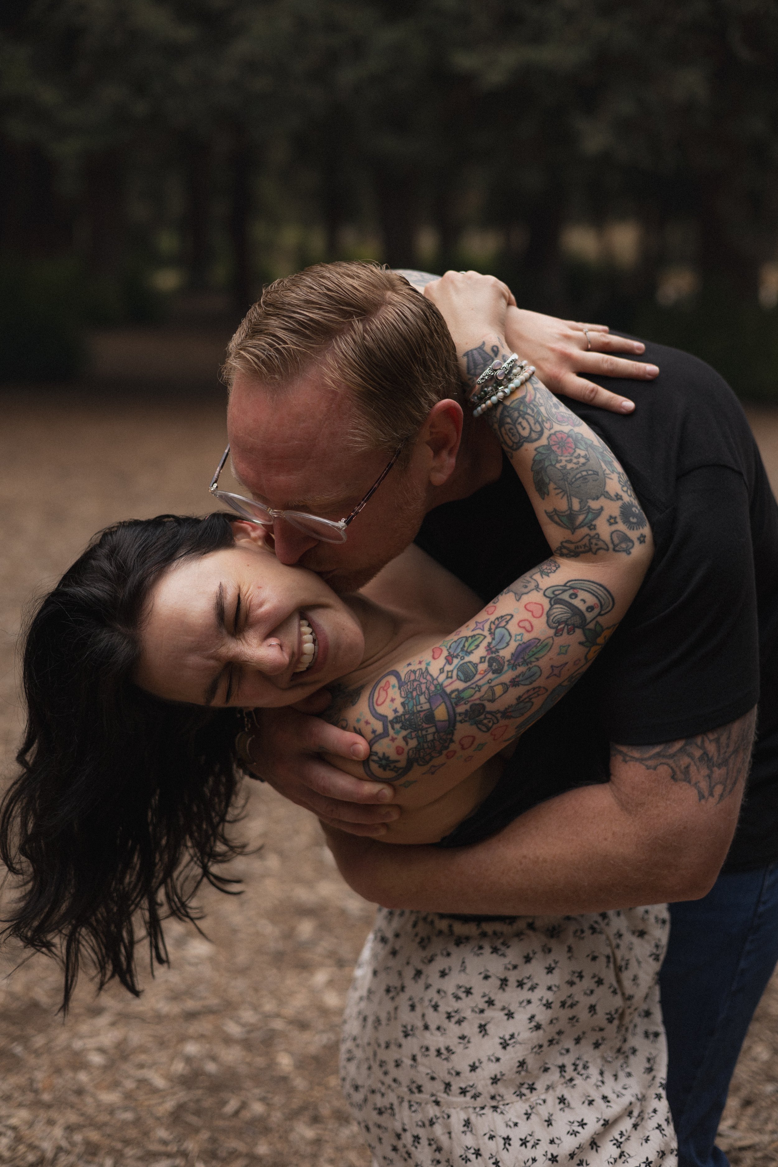 A couple dancing outdoors with a young girl in a white dress with black star patterns. The man, with glasses and tattoos on his arms, is kissing the woman's forehead while she smiles and has her eyes closed. The background is blurred with trees and d