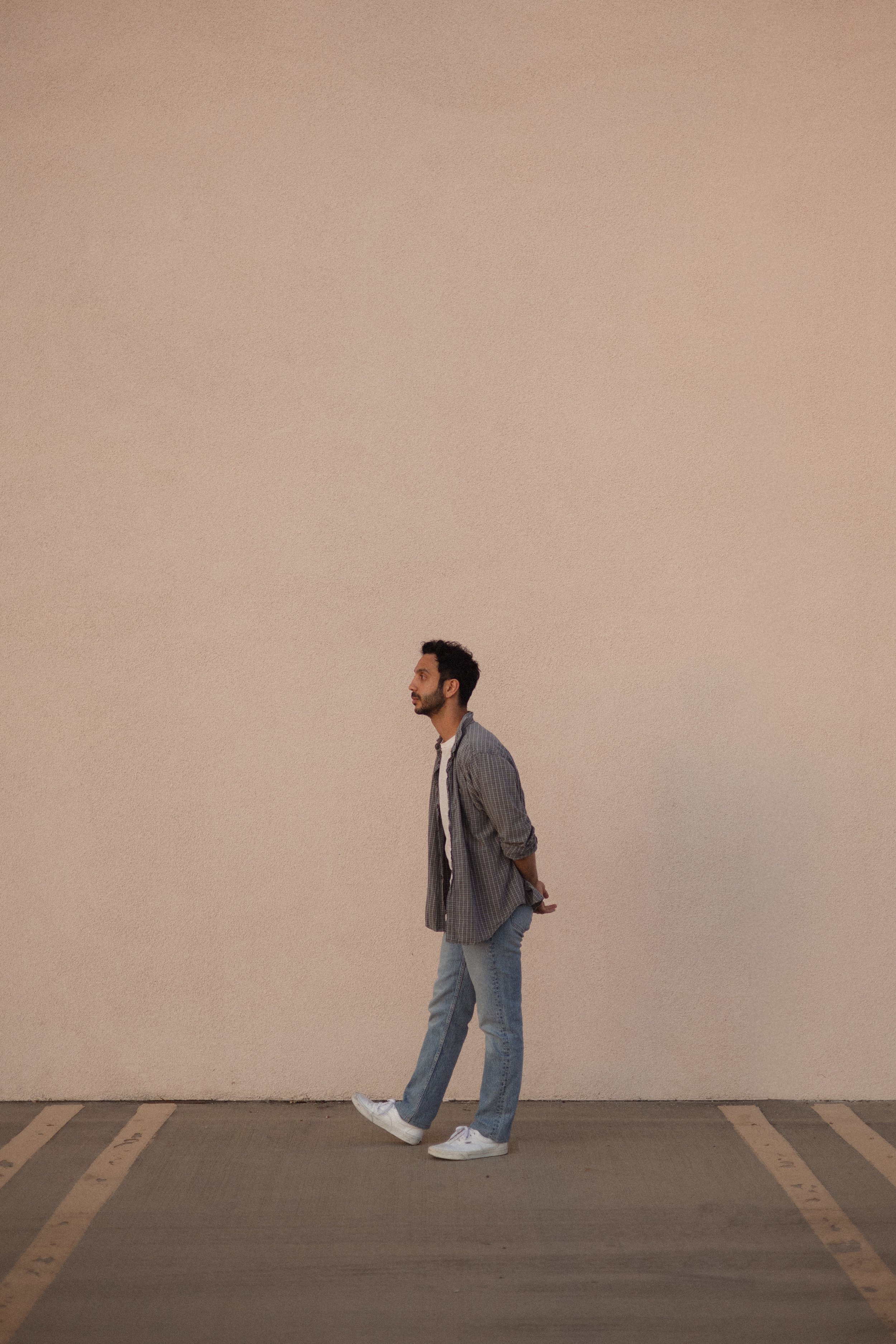 A young man with a beard in a gray plaid shirt and blue jeans walking in a parking lot with marked parking spaces against a plain beige wall.