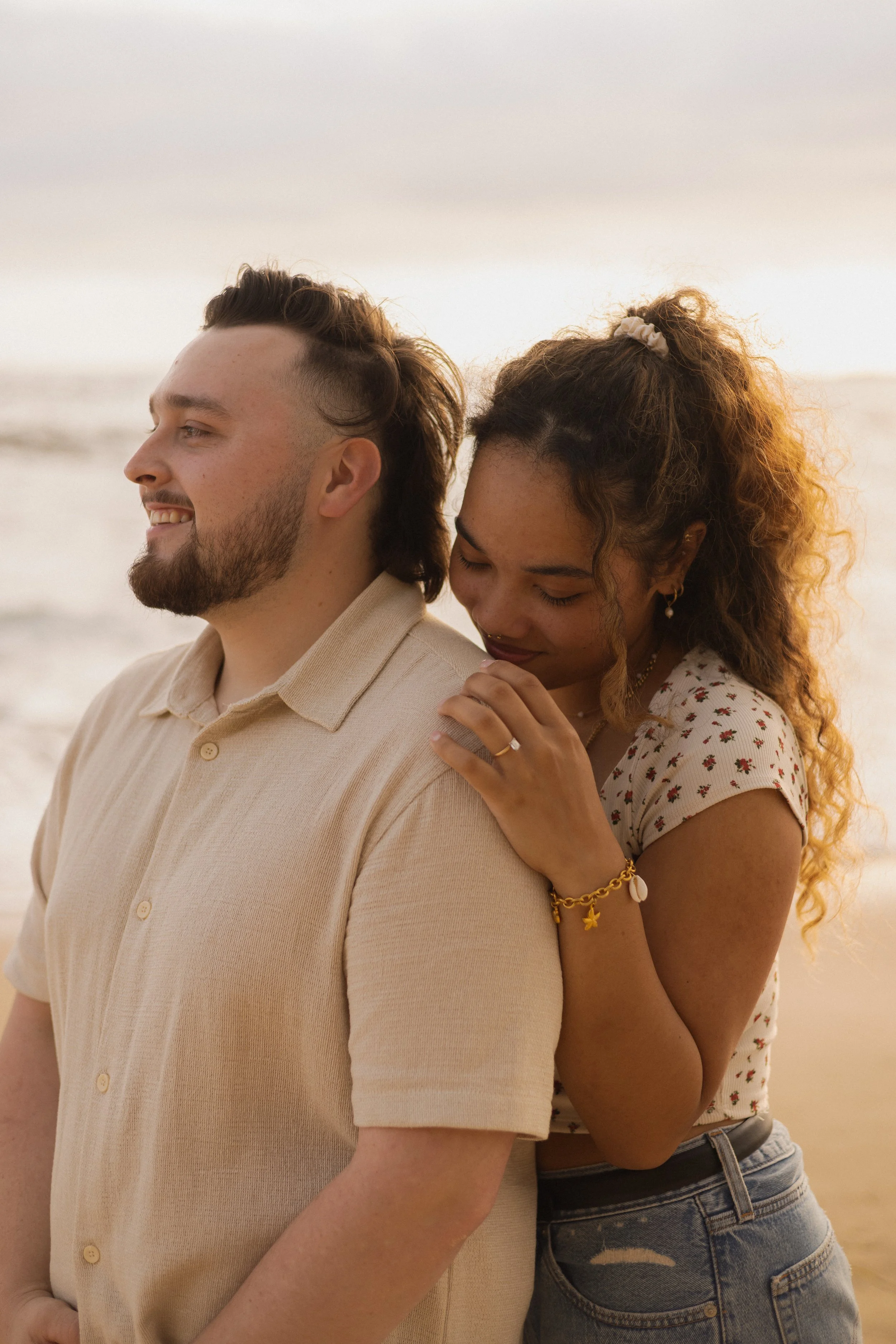 A smiling man wearing a beige shirt stands in front of a woman with curly hair tied up with a scrunchie, who is hugging him from behind. They are at the beach during sunset.