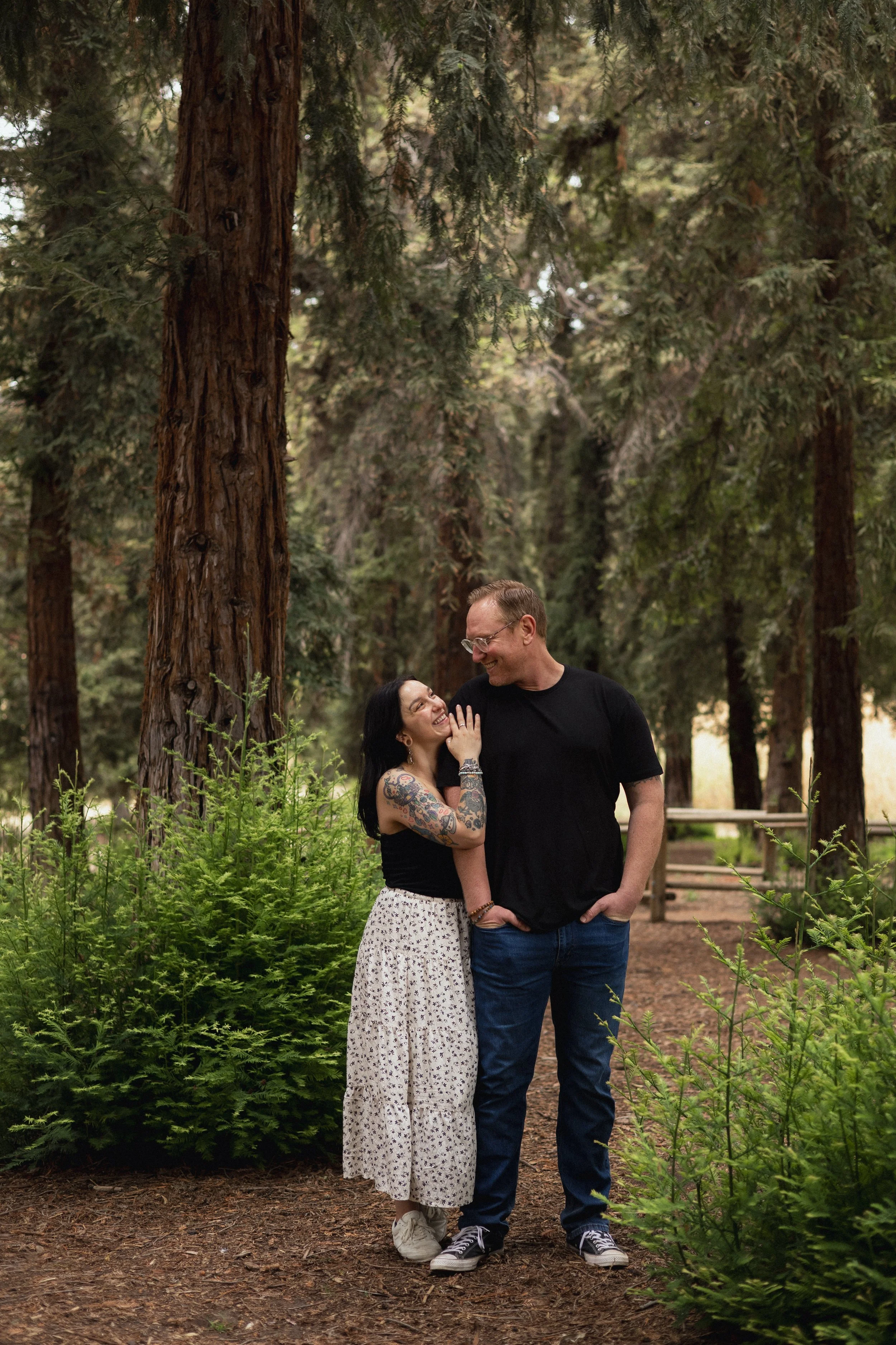 A smiling couple stands together in a forest. The woman has tattoos on her arms and is looking up at the man, who is looking down at her. The woman wears a white skirt with black patterns and a black sleeveless top, while the man is dressed in a black t-shirt and blue jeans. They are standing close, surrounded by tall trees and lush green foliage.