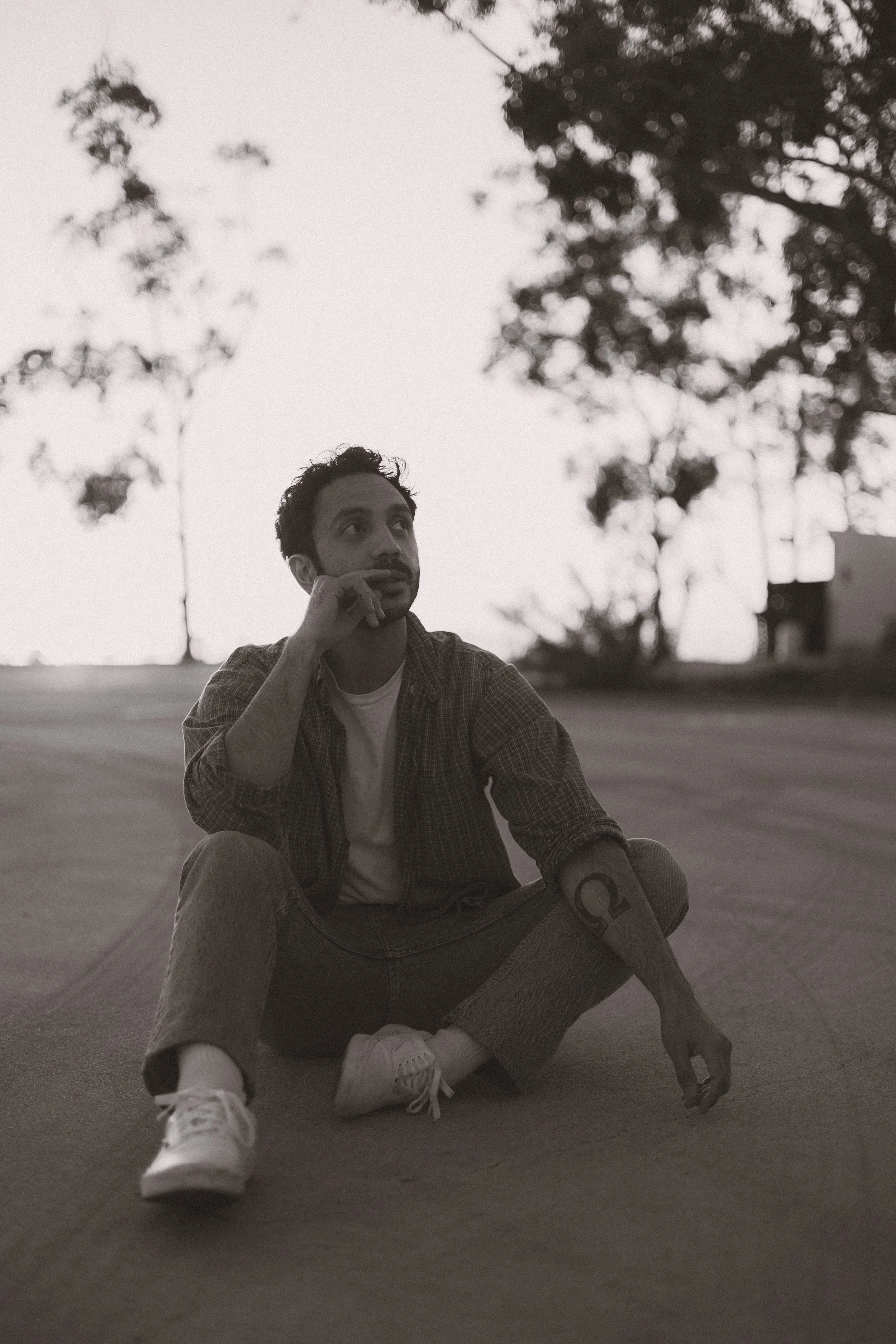 A man sitting on the ground outdoors in black and white, with trees and a building in the background, wearing casual clothing and sneakers, looking thoughtful.
