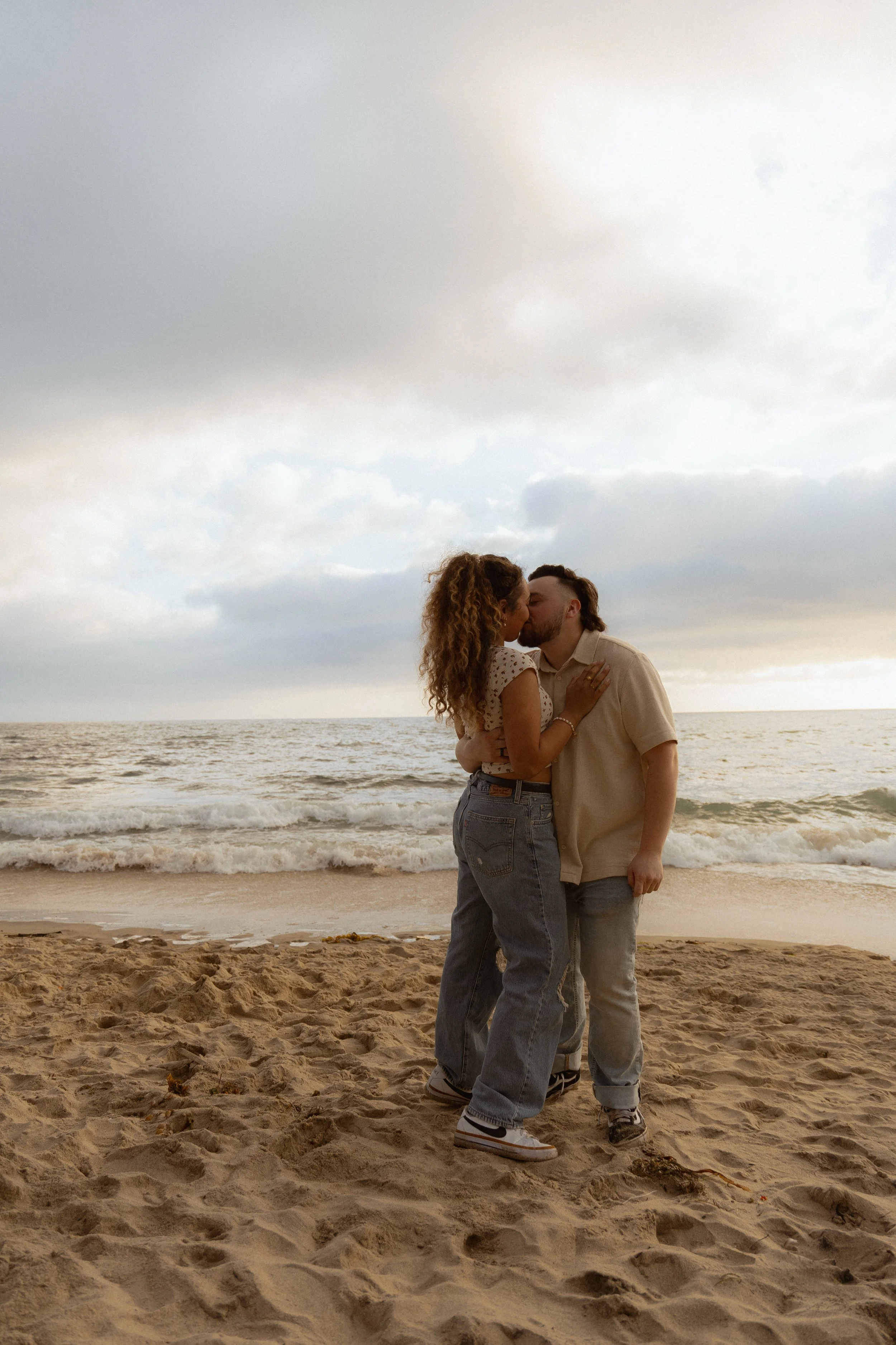 A couple kissing on a beach with sand, ocean waves, and a cloudy sky in the background.
