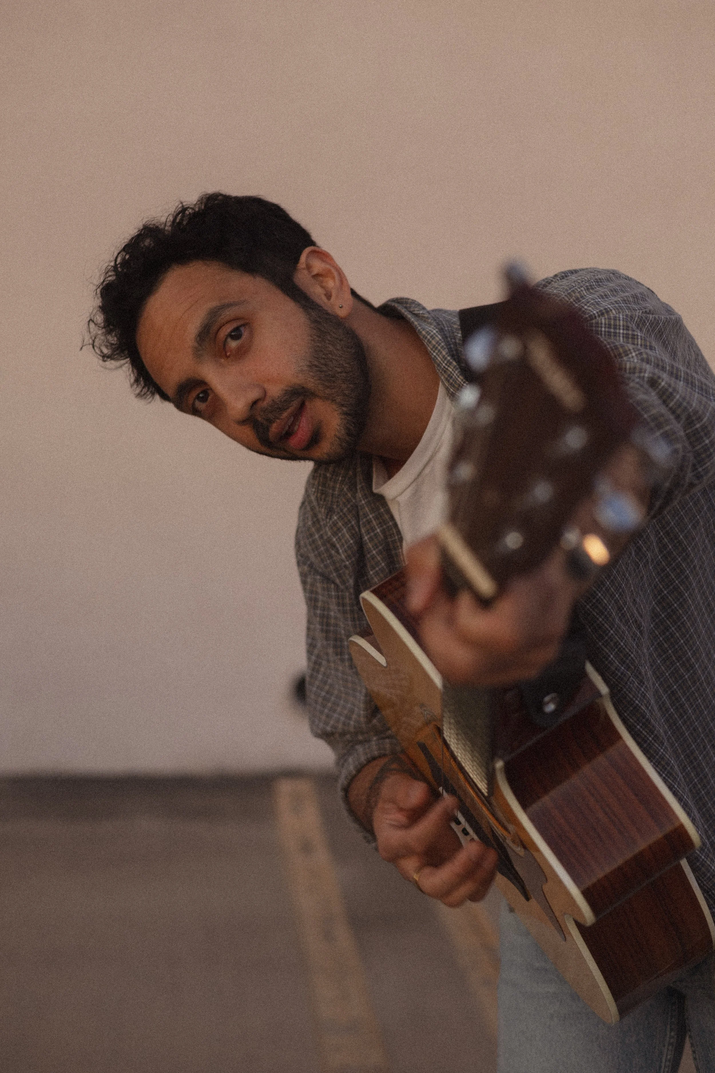 A man with dark hair and a beard, wearing a checkered shirt and earring, is strumming and playing an acoustic guitar.