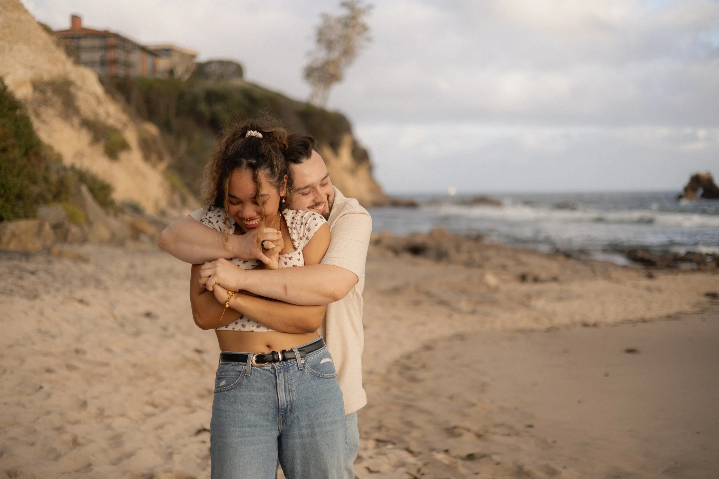 A couple hugging on a beach, with houses on a hill in the background and the ocean waves nearby.