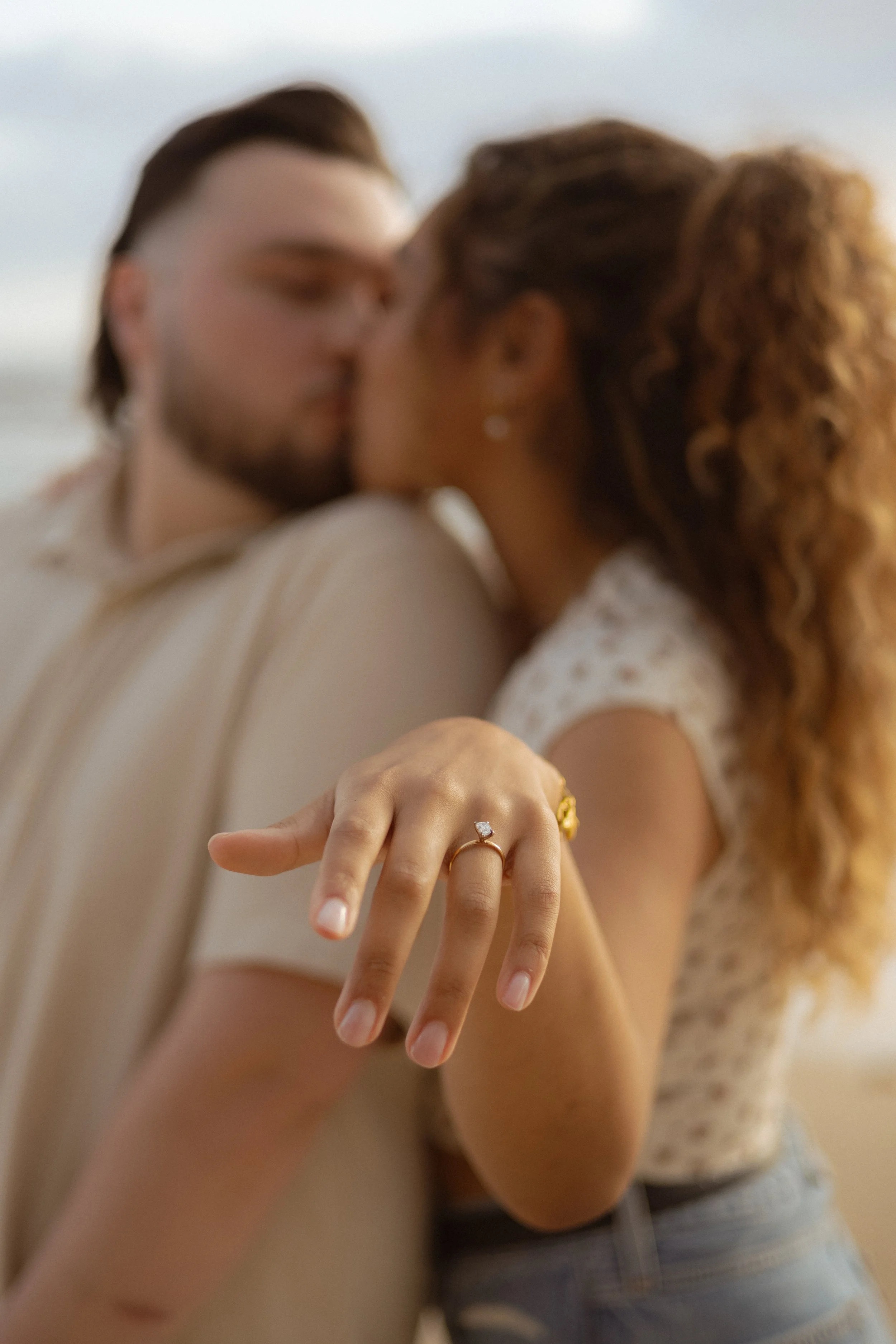 Close-up of a woman's hand displaying an engagement ring and wedding band. In the background, a couple is about to kiss, standing outdoors with a blurred sky and water scene.