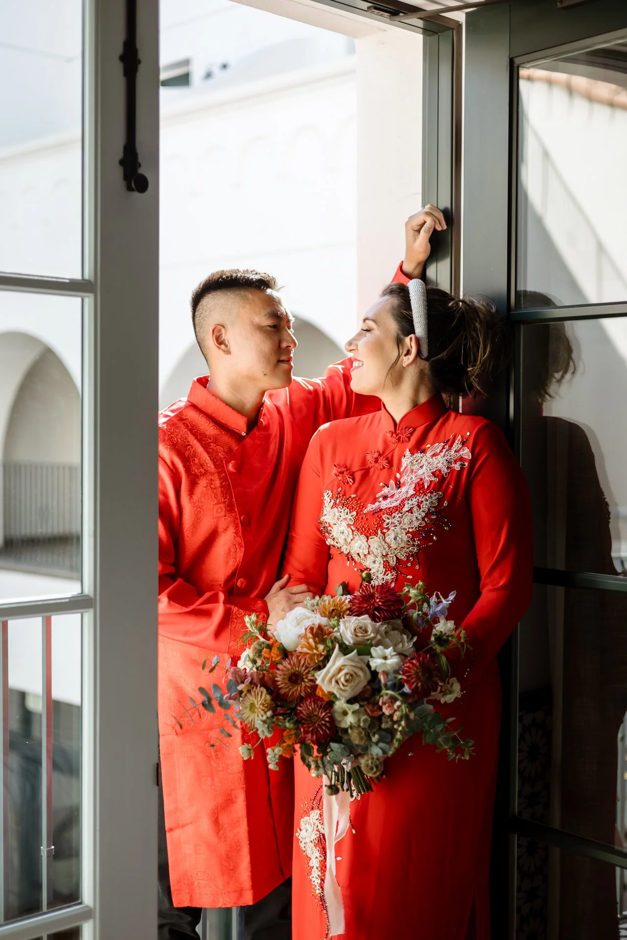 An Asian man and a Caucasian woman in traditional red Chinese wedding attire standing close together, with the man leaning into the woman near a doorway, holding a bouquet of flowers, in a brightly lit indoor setting.