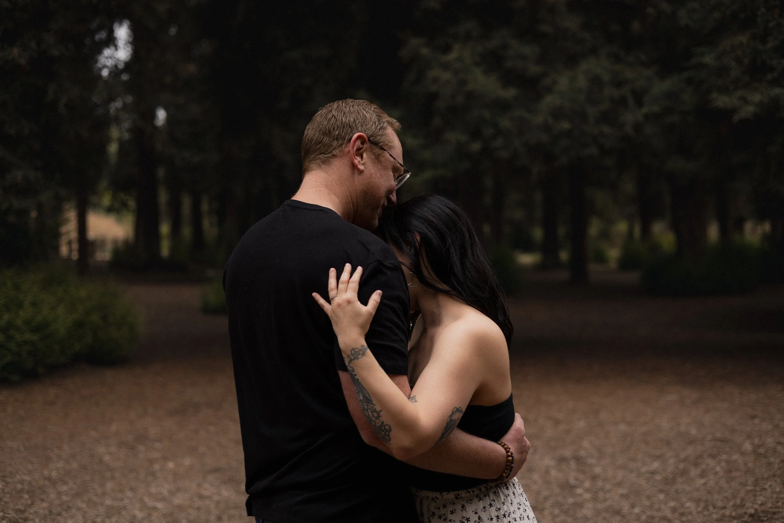 A man and woman hugging outdoors during dusk or evening, with a blurred background of trees and a park.