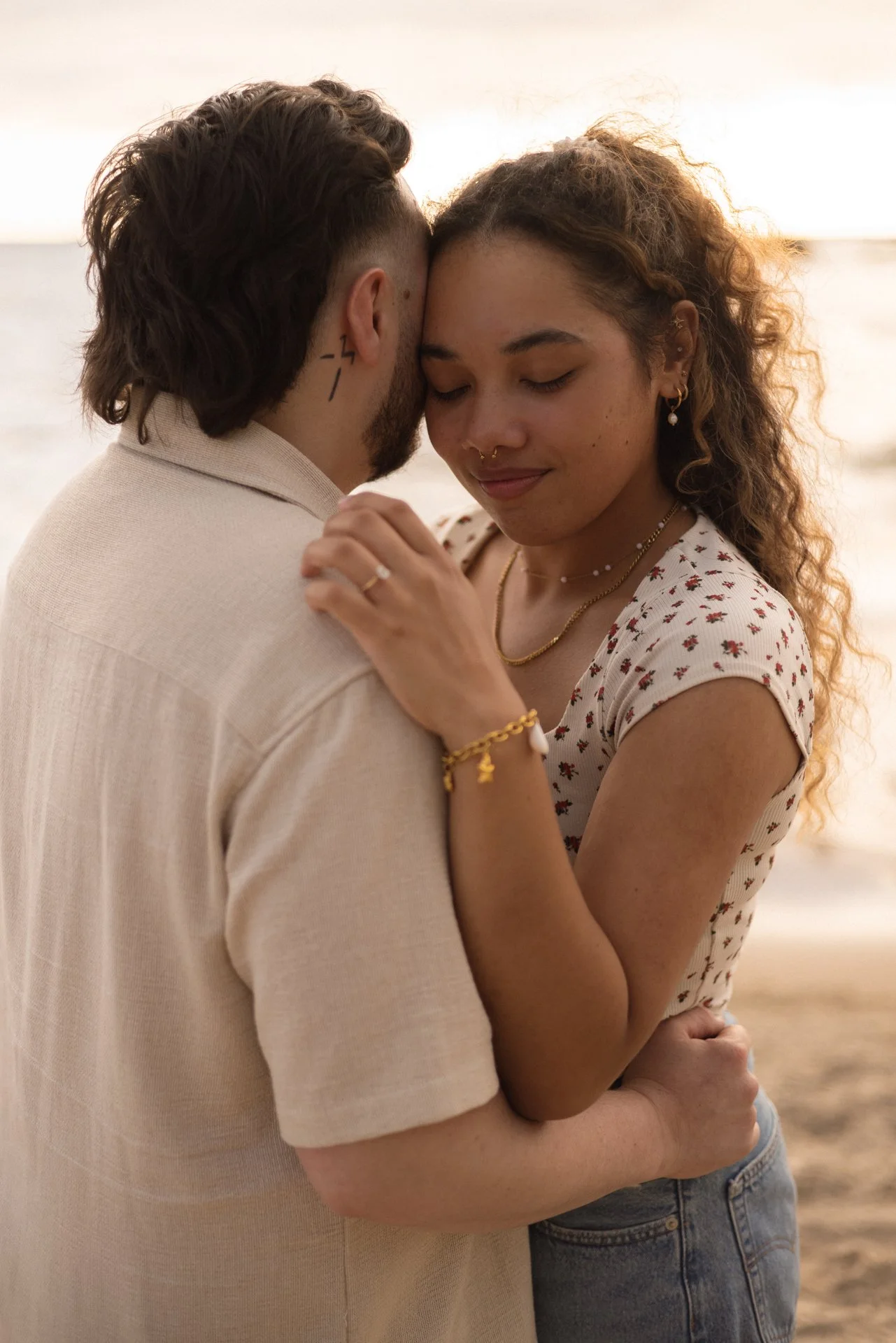 A man and woman embrace on a beach at sunset, with their foreheads touching and eyes closed, conveying affection and intimacy.