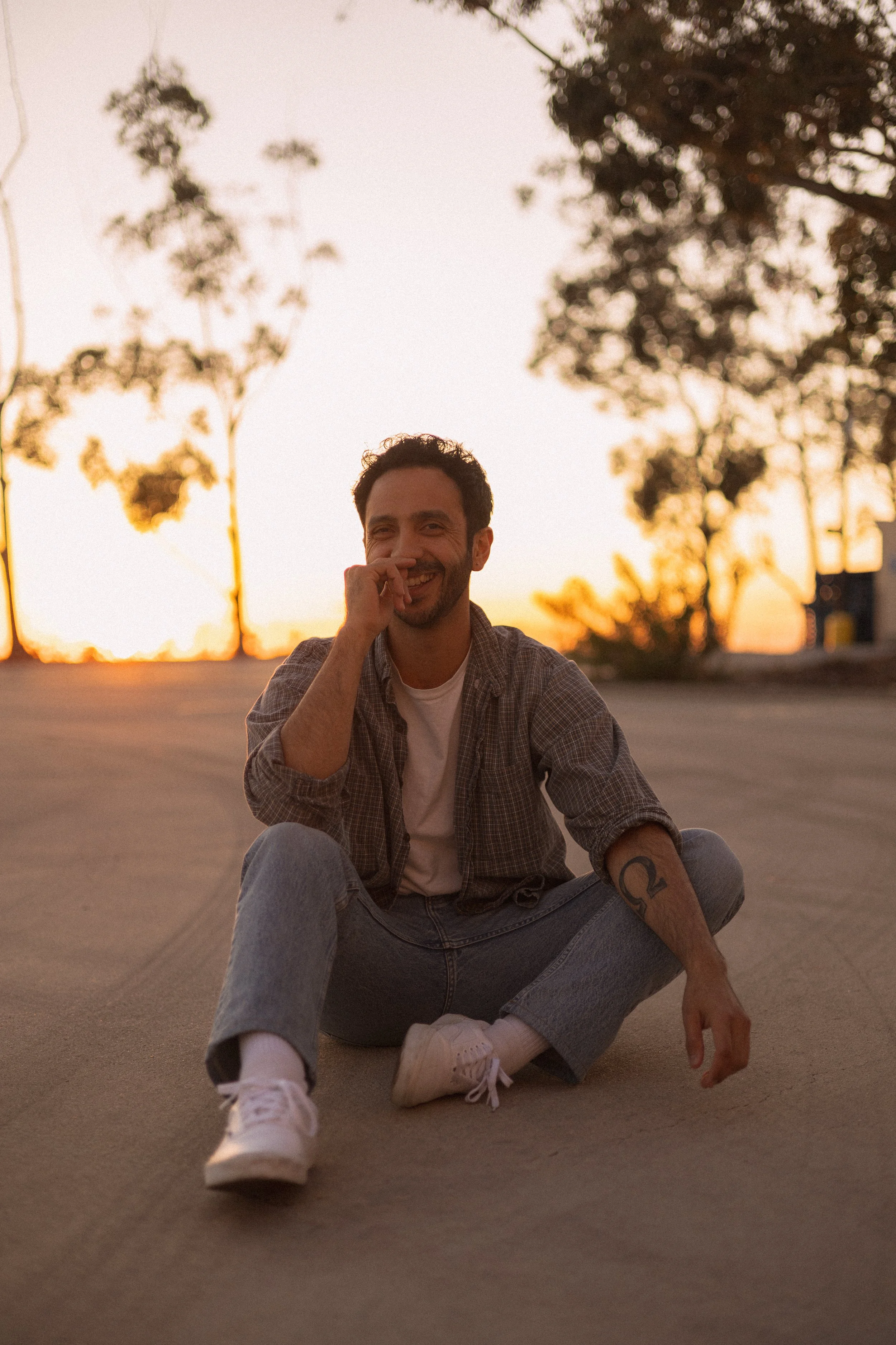 A man sitting on the ground outdoors during sunset, smiling and holding his nose. He has dark curly hair, a tattoo on his left forearm, and is dressed in a checkered shirt, white t-shirt, and jeans. There are trees and a clear sky in the background.