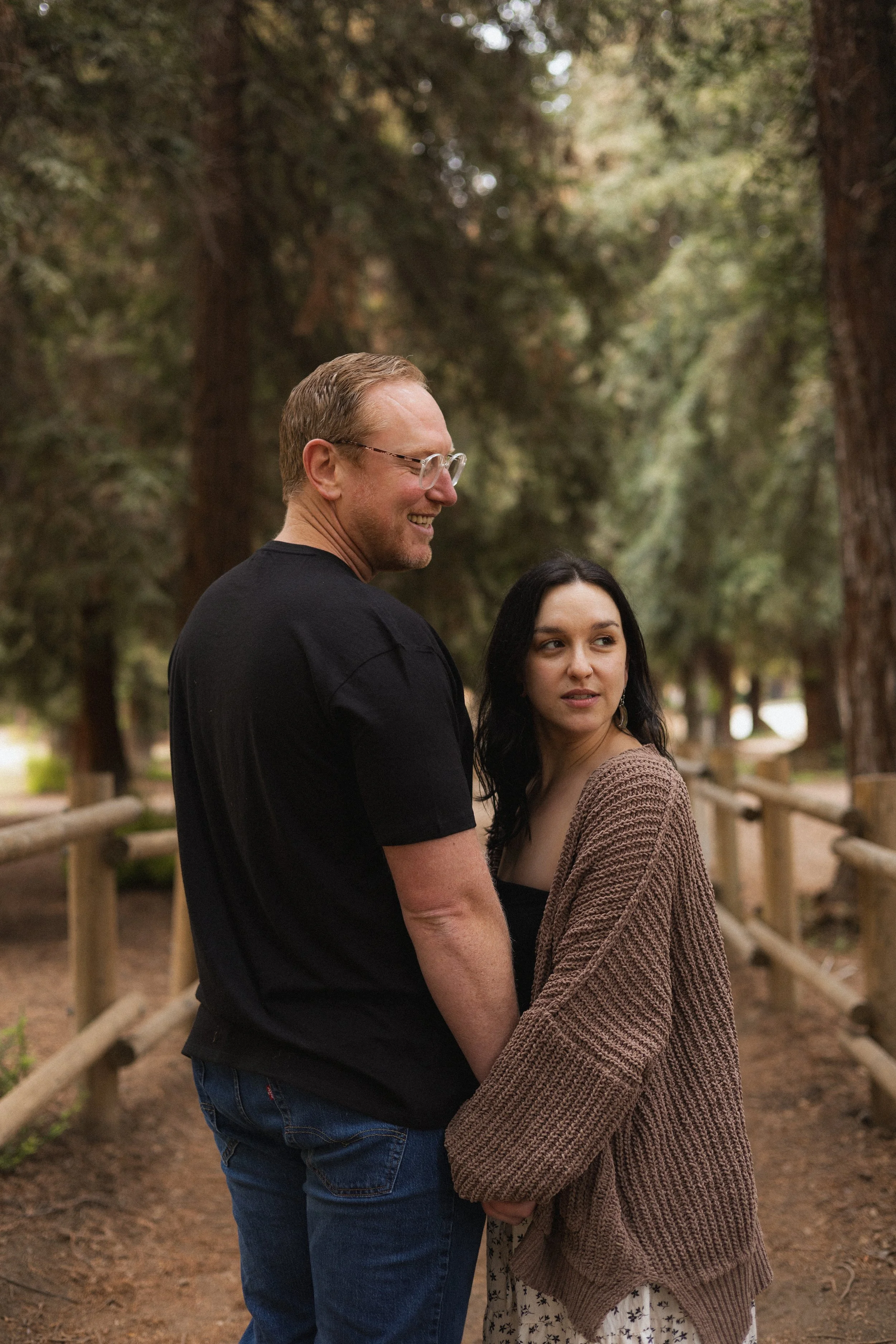 A smiling man and woman standing outside in a wooded area, close together, with the man looking at the woman and the woman looking slightly away. They are near a wooden fence, and the background shows tall trees with greenery.