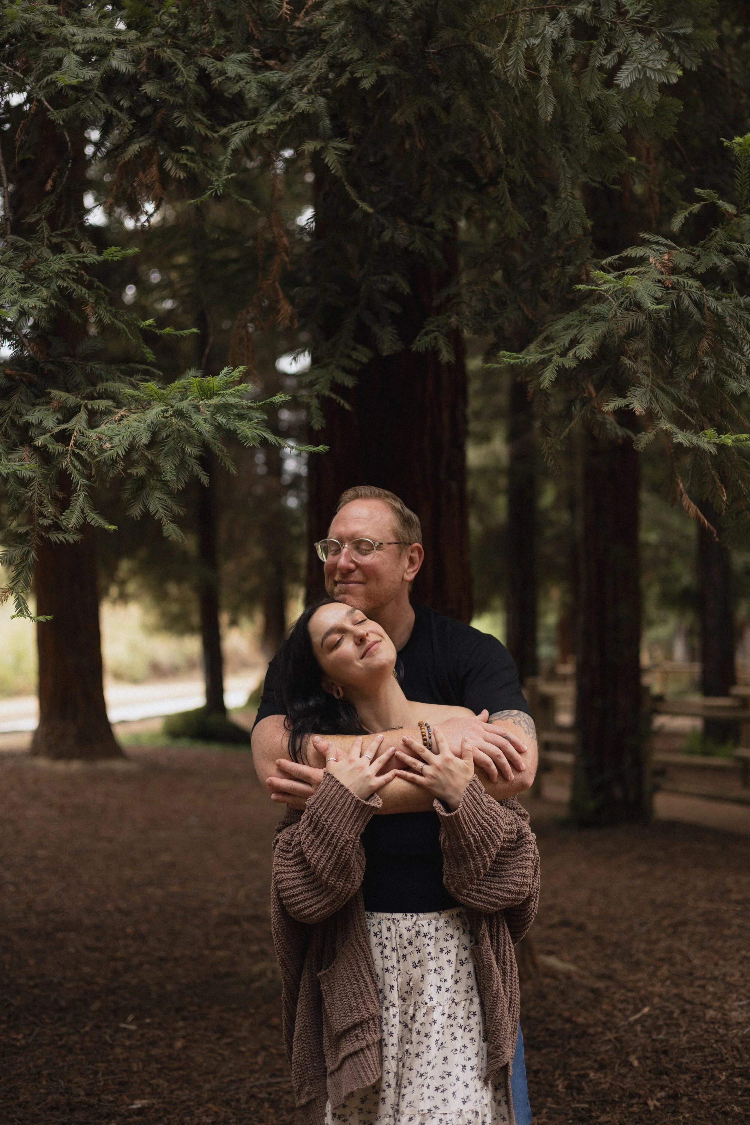 A man and a woman embrace in a forested park, surrounded by tall trees with green foliage. The woman is resting her head on the man's chest with a content expression, while the man has a gentle smile and closes his eyes. The scene appears peaceful an