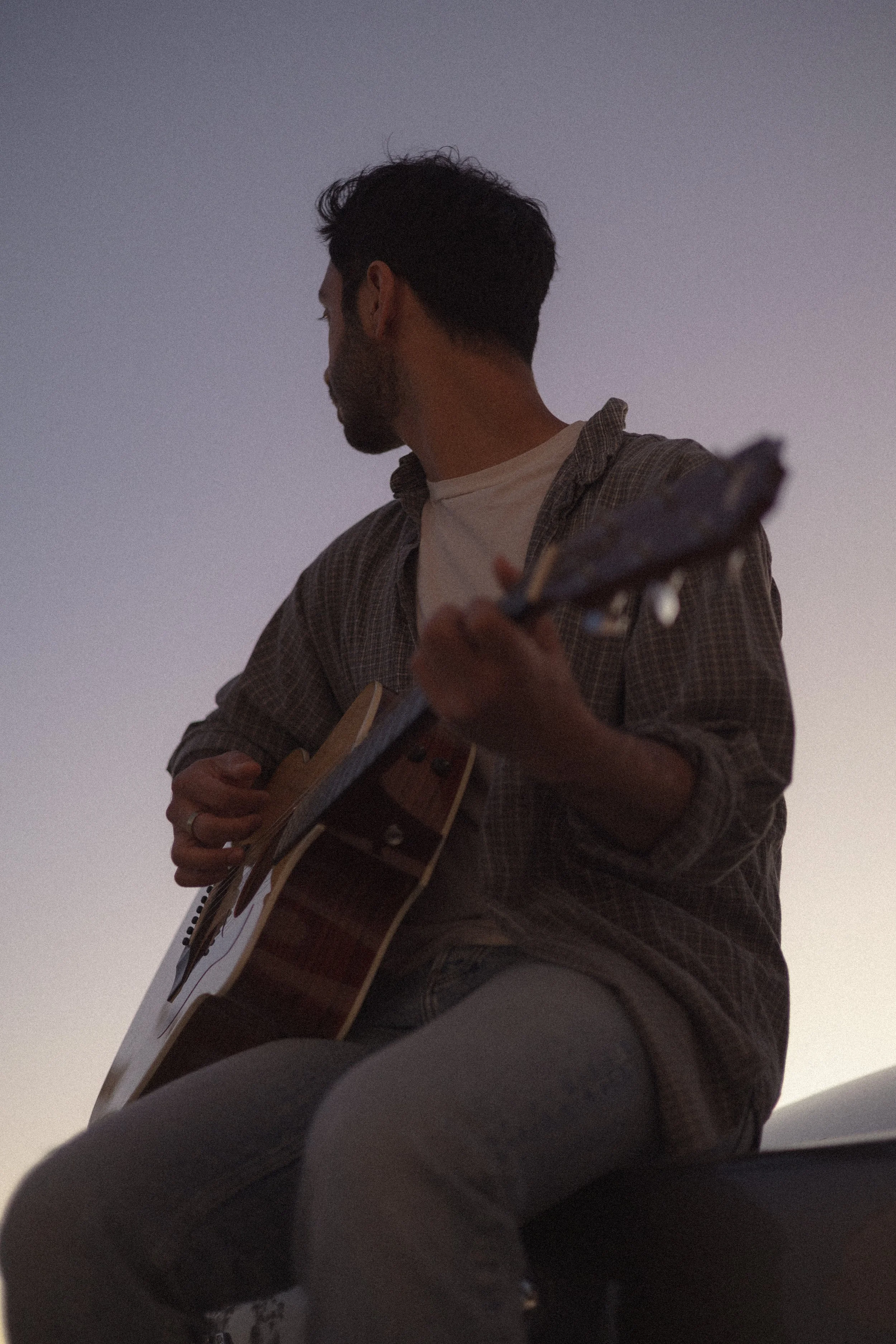 A man sitting outdoors playing an acoustic guitar during dusk or dawn.