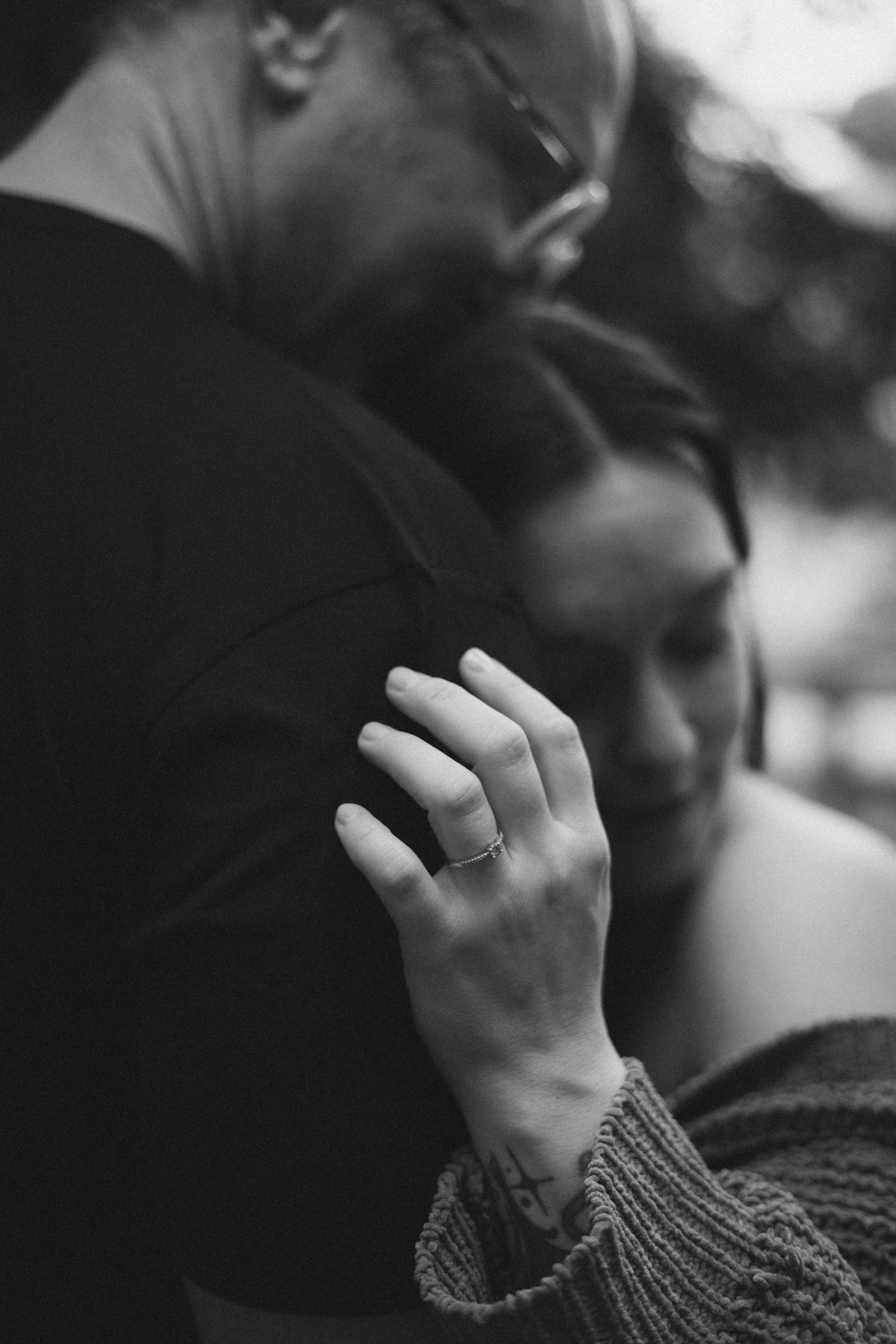 A close-up black and white photo of a person's hand resting on another person's shoulder, with part of their face visible, showing glasses and long hair.