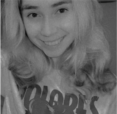 Black and white image of a person smiling, wearing a "Wolverine's" T-shirt with long hair.