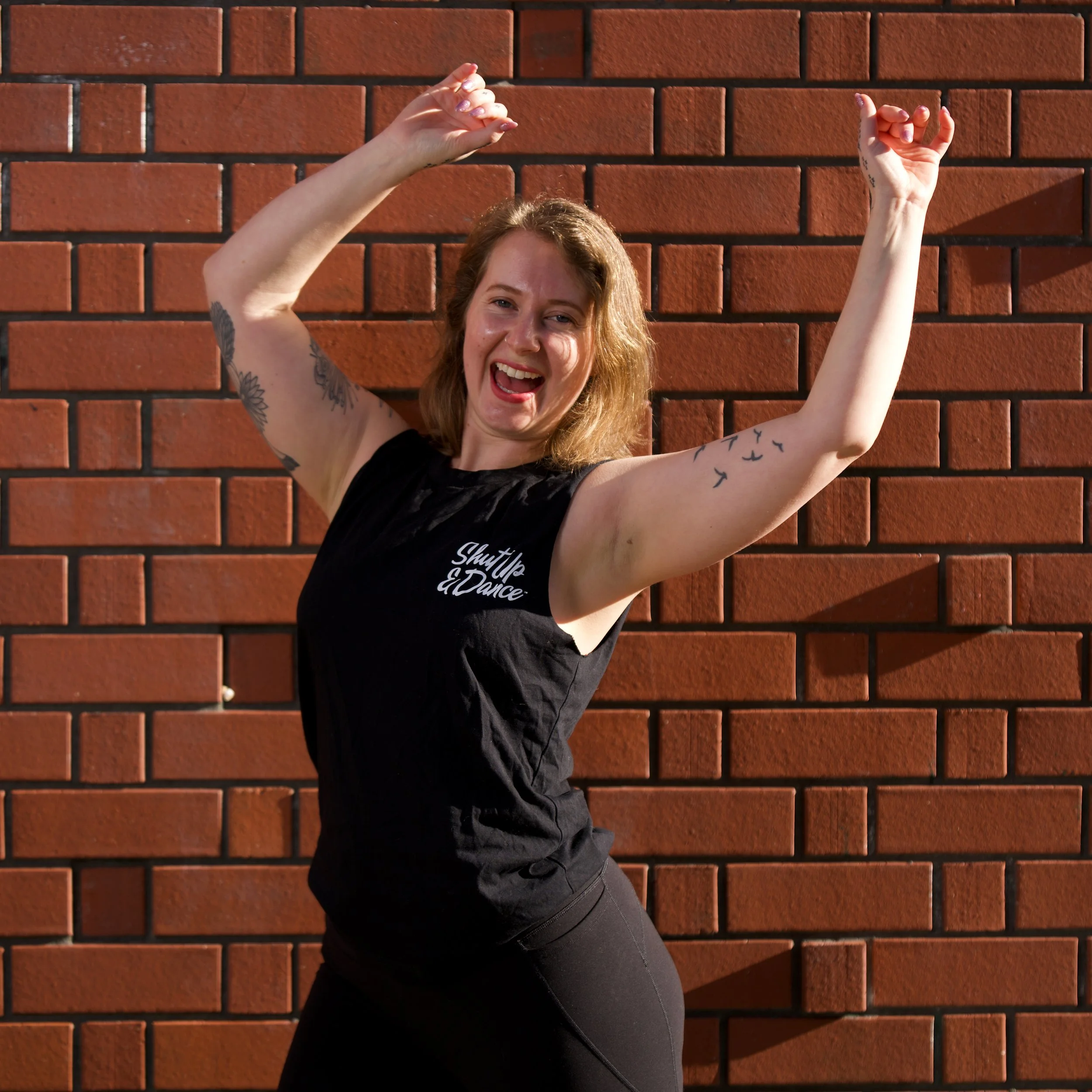 Gloria dances with a smile on her face wearing a black shut up and dance t-shirt, with black leggings. The background is a brick wall, the sun is shining