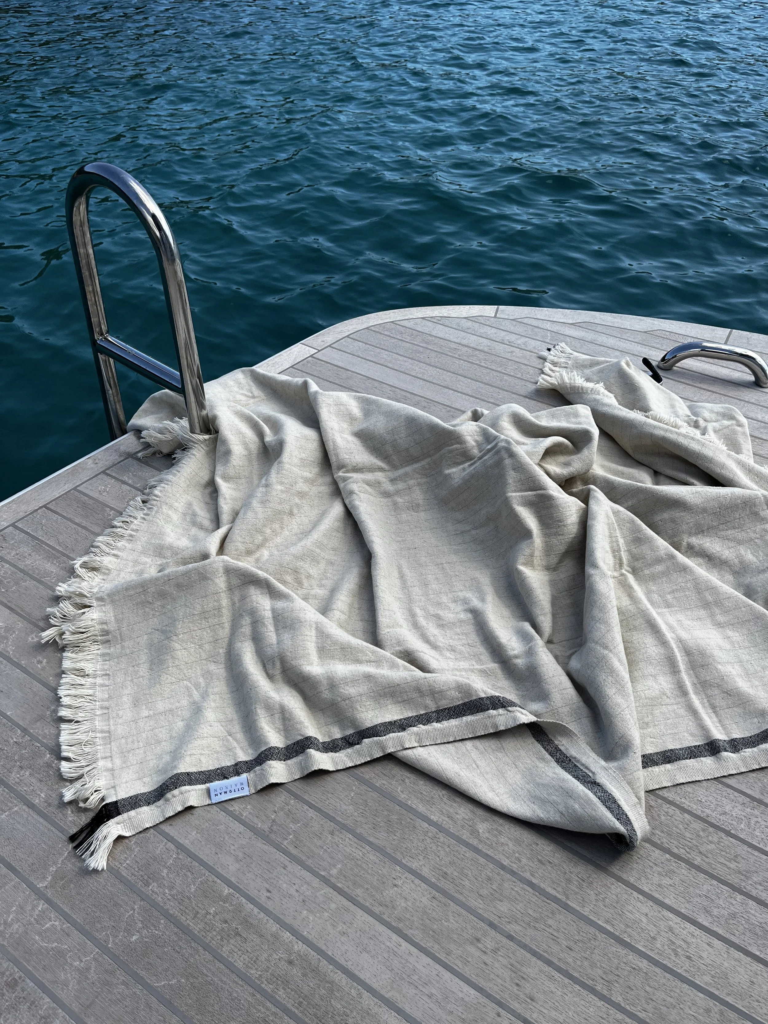 A beige and black contemporary striped blanket on the wooden deck of a yacht, with a metal ladder leading into the sea, and a white towel with fringes on the blanket.  It represents costal, outdoor living.