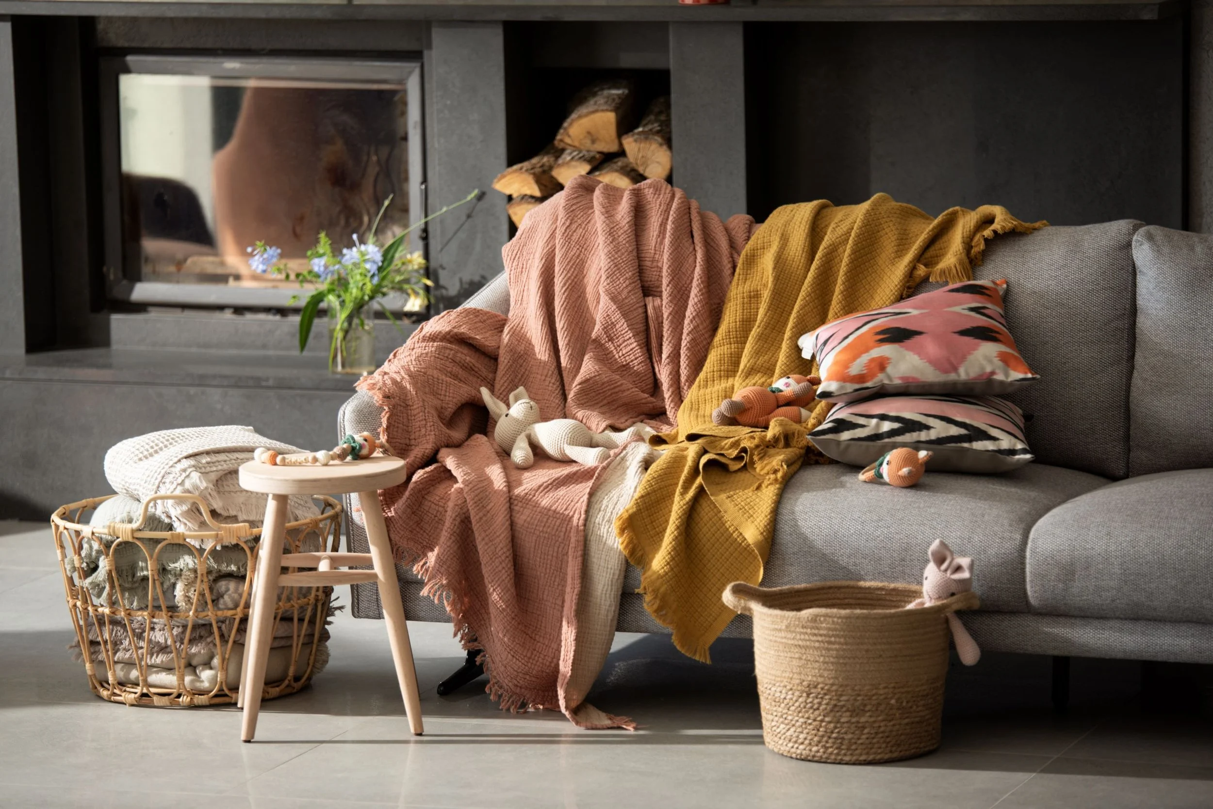 A living room with a sofa draped with pink and mustard woven blankets, a colourful pillow, a wicker basket with plush toys and a small round wooden table. It depicts a cosy home with premium interiors.