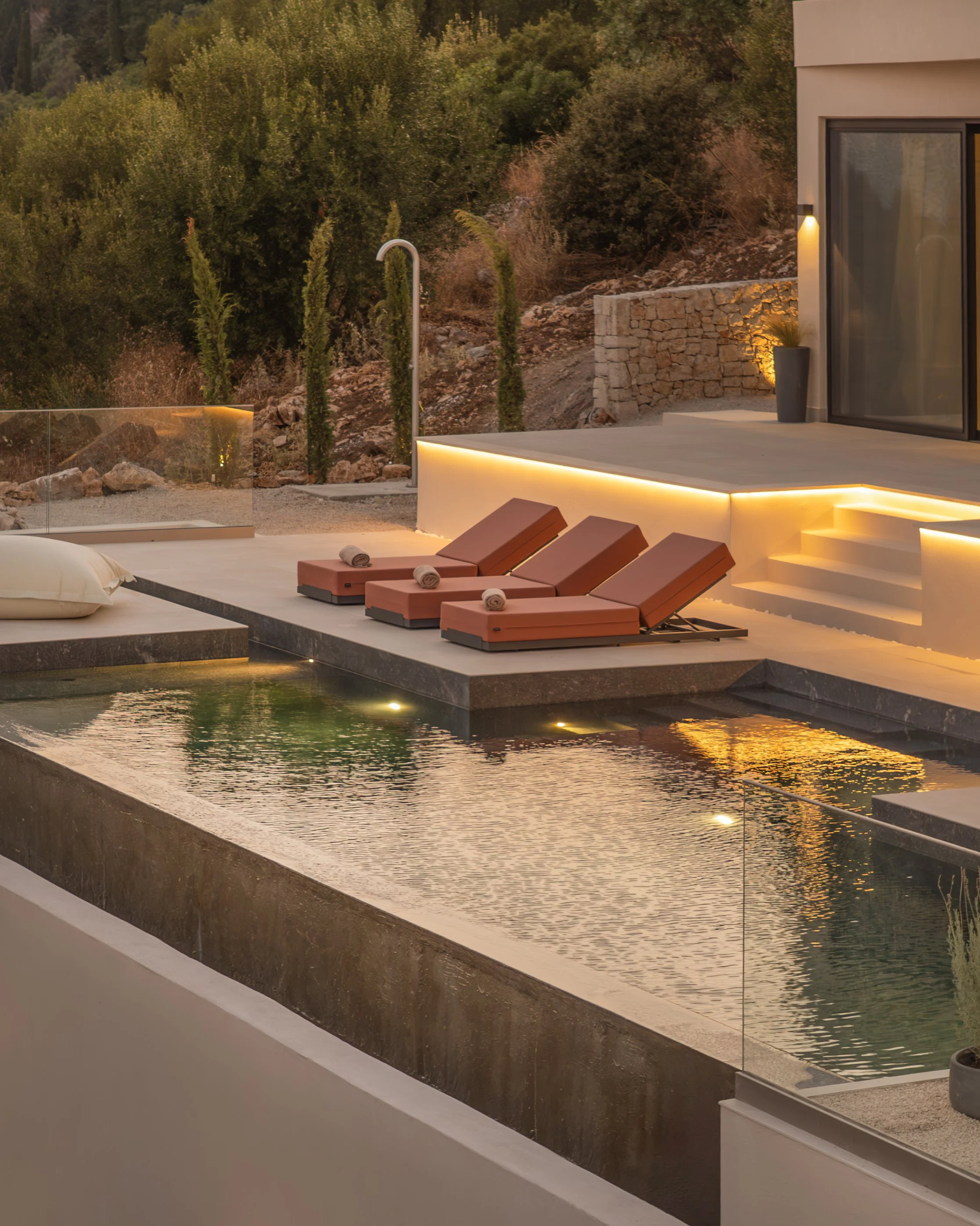 Outdoor pool area with three brown lounge chairs, rolled towels, and a pool with underwater lights, surrounded by a modern patio with steps and a glass railing, set against a natural hillside with trees and bushes at sunset.