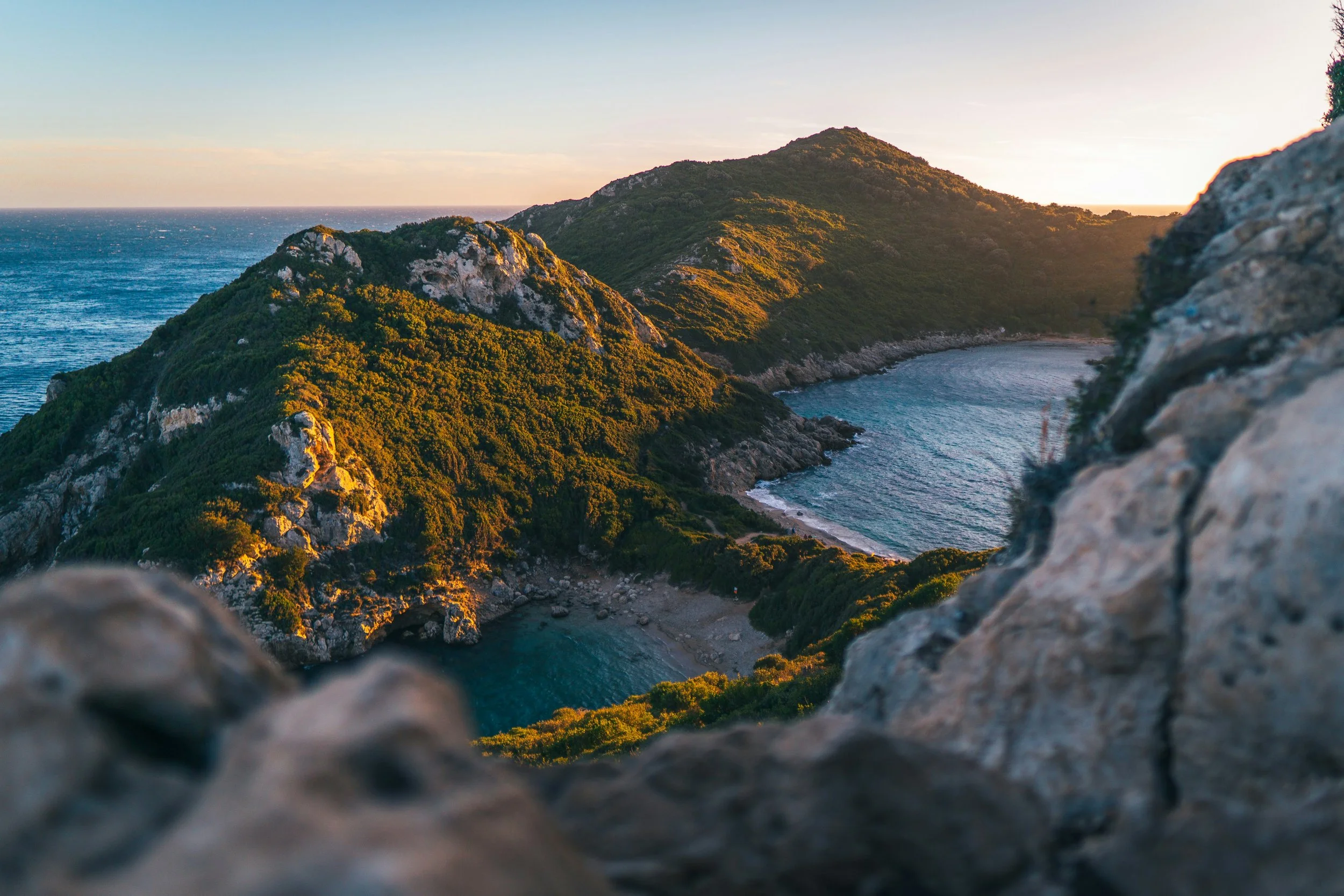 Scenic view of a coastal bay with green hills and rocky coastline, taken at sunrise or sunset with soft lighting.
