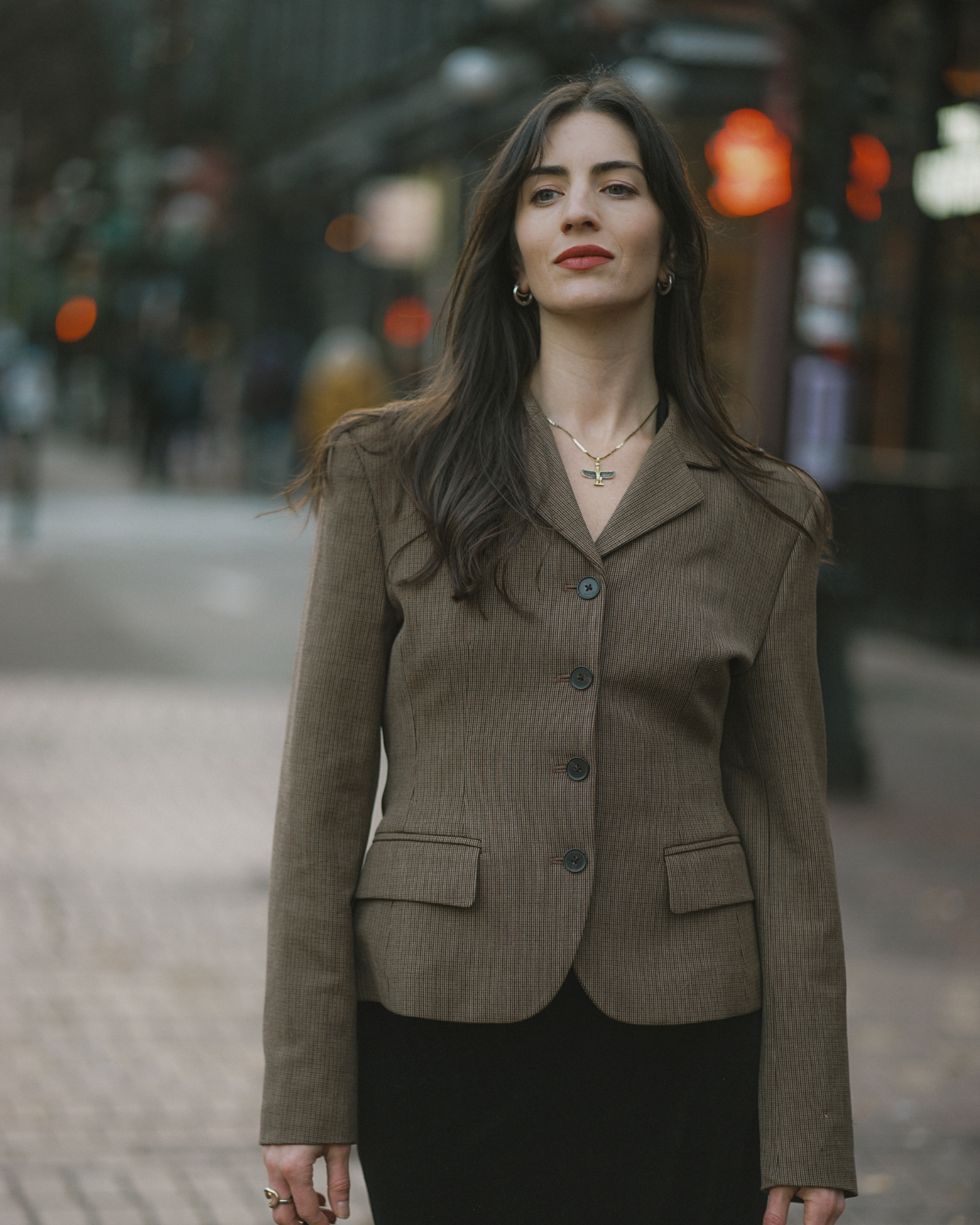 A woman with long dark hair wearing a brown blazer, black top, and jewelry standing on city street with blurred lights in background.