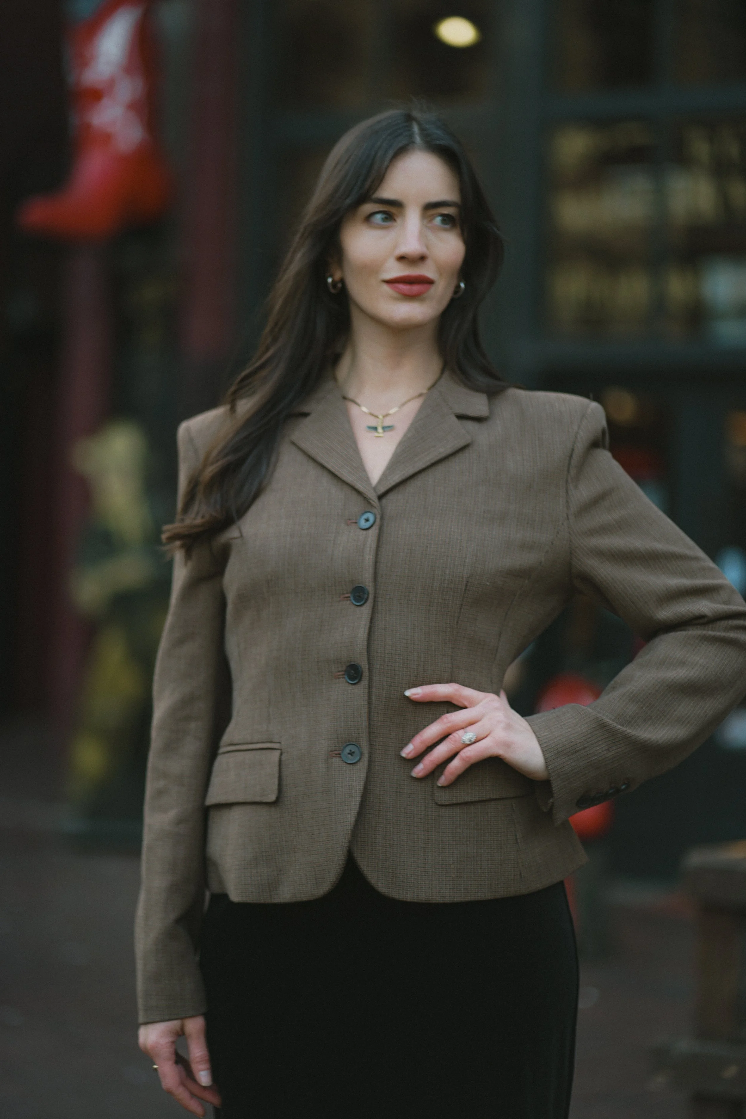 A woman with long dark hair, wearing a brown blazer and a black skirt, standing outdoors in front of a building with glass windows and red decorations.