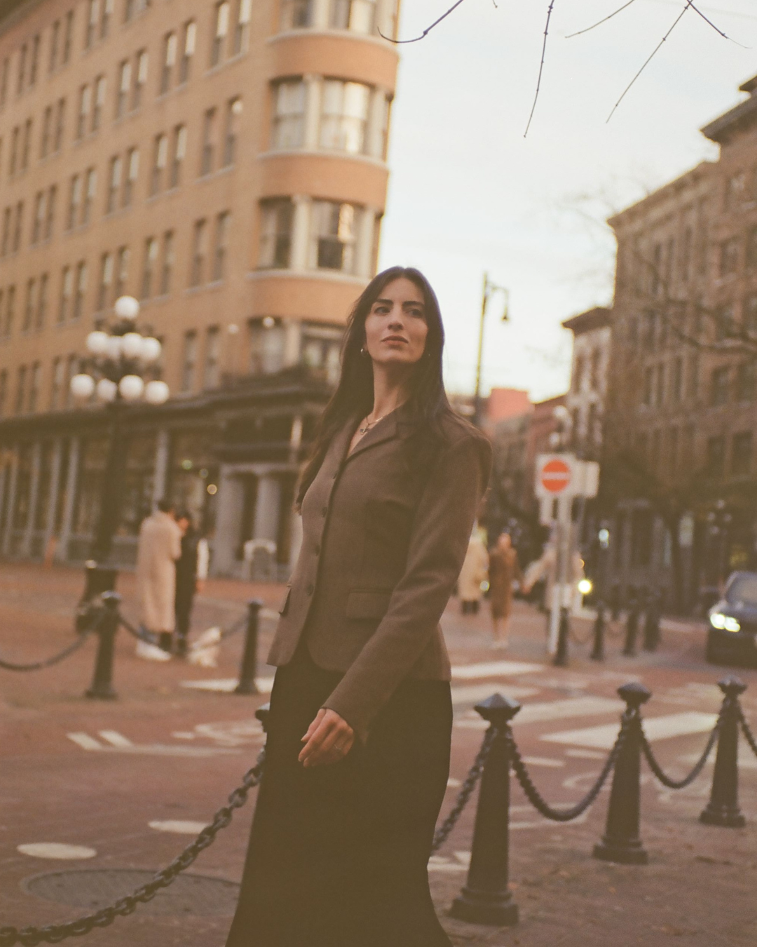 A woman standing on a city street at sunset, wearing a brown blazer and black skirt, with old multi-story buildings and street lamps in the background.