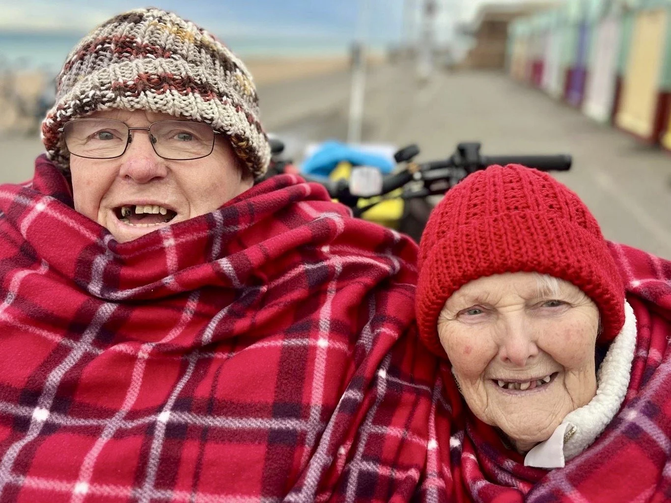 Two participants smiling at the camera whilst sit in the front of a trishaw. They are wrapped up in checked blankets and wearing woolly hats. There are beach huts and the sea in the background