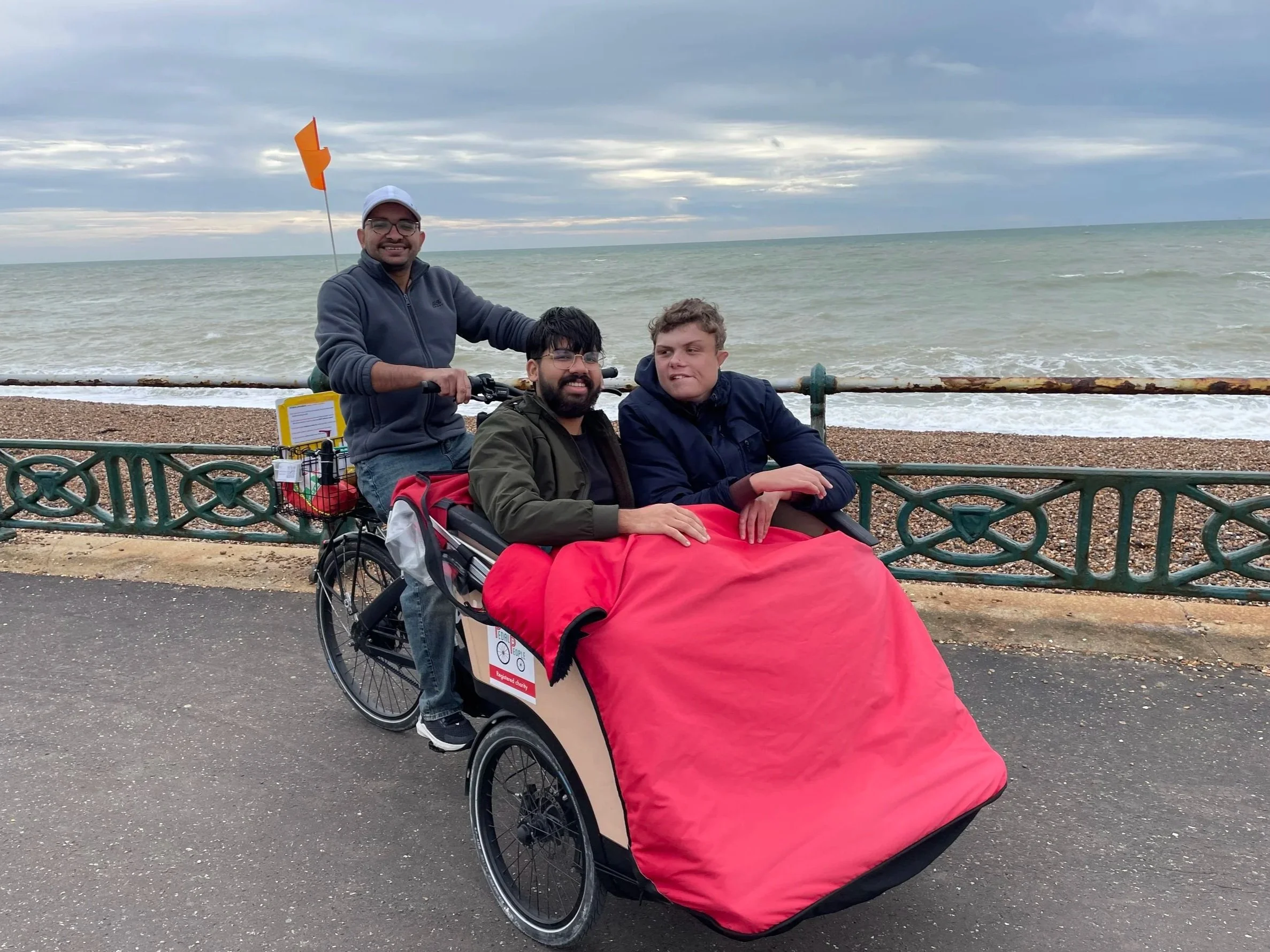 Two participants sit in the front of a bright red trishaw with the Cycle-Pilot sitting behind them. They are on a seafront promenade with a ornate green fence behind them. The beach, sea and cloudy sky can be seen in the background.