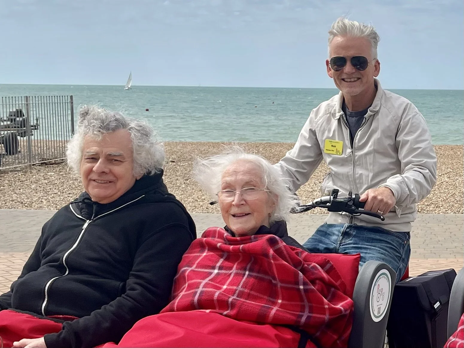 Two people wrapped in blankets sit at the front of a trike, smiling, with a rider behind them on the seafront