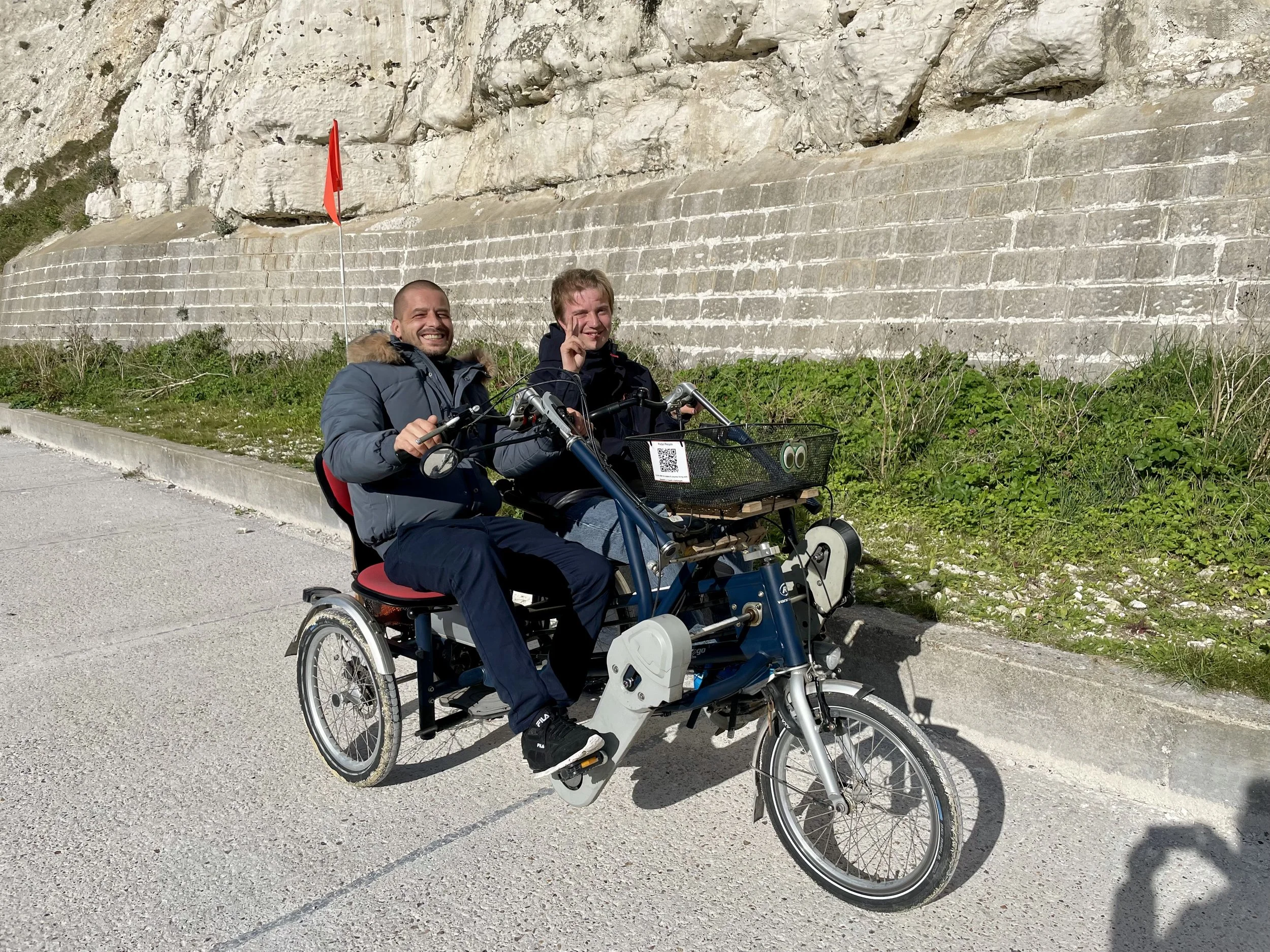 Two participants sit on a side-by-side tandem. They are smiling at the camera and one is doing a 'peace' hand sign. They are in front of a white chalk cliff. 