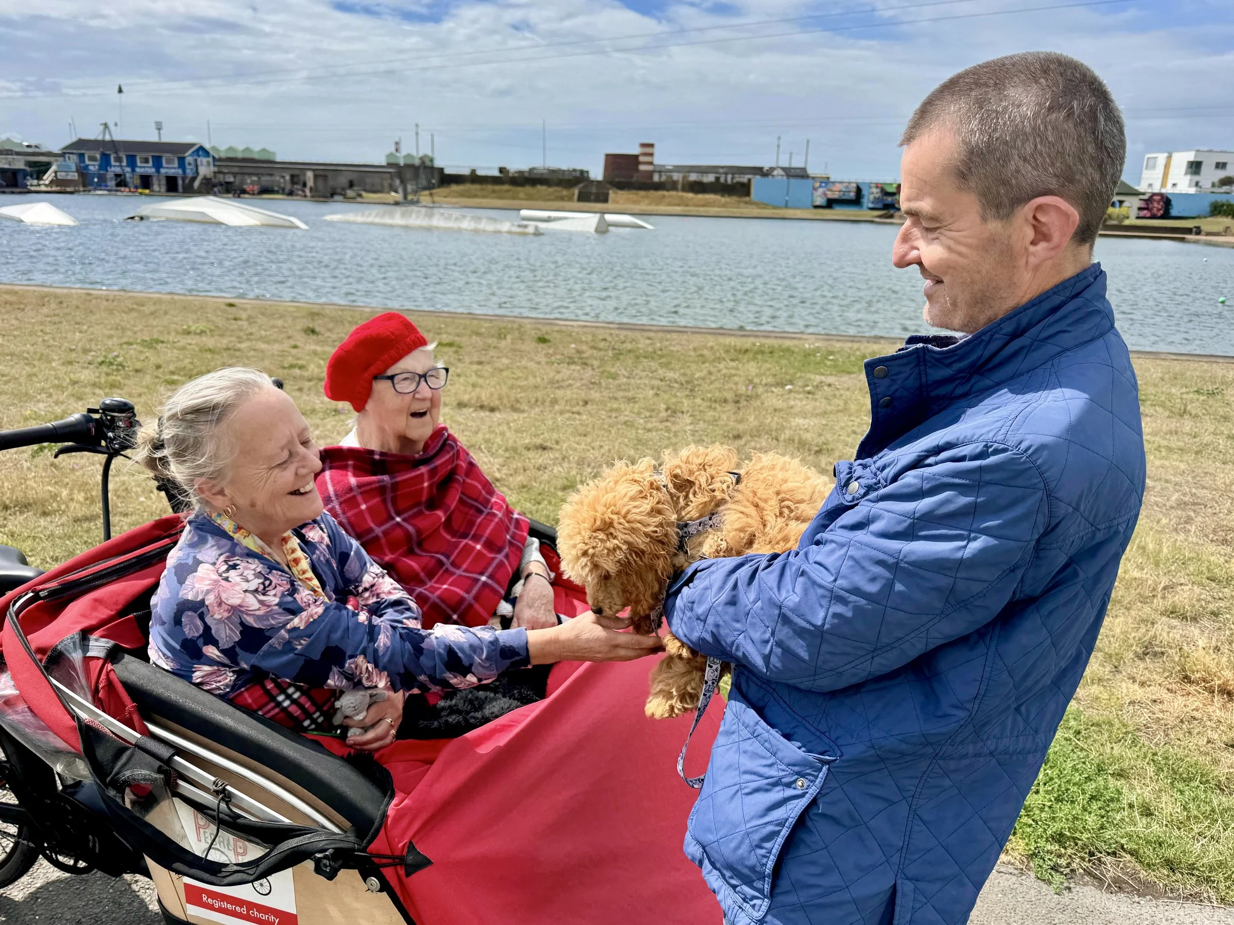 Two women, wrapped in blankets, sit in the front carriage of a trishaw cycle. They are petting a small auburn dog, who is being held up to them by a smiling man. Hove Lagoon is in the background.