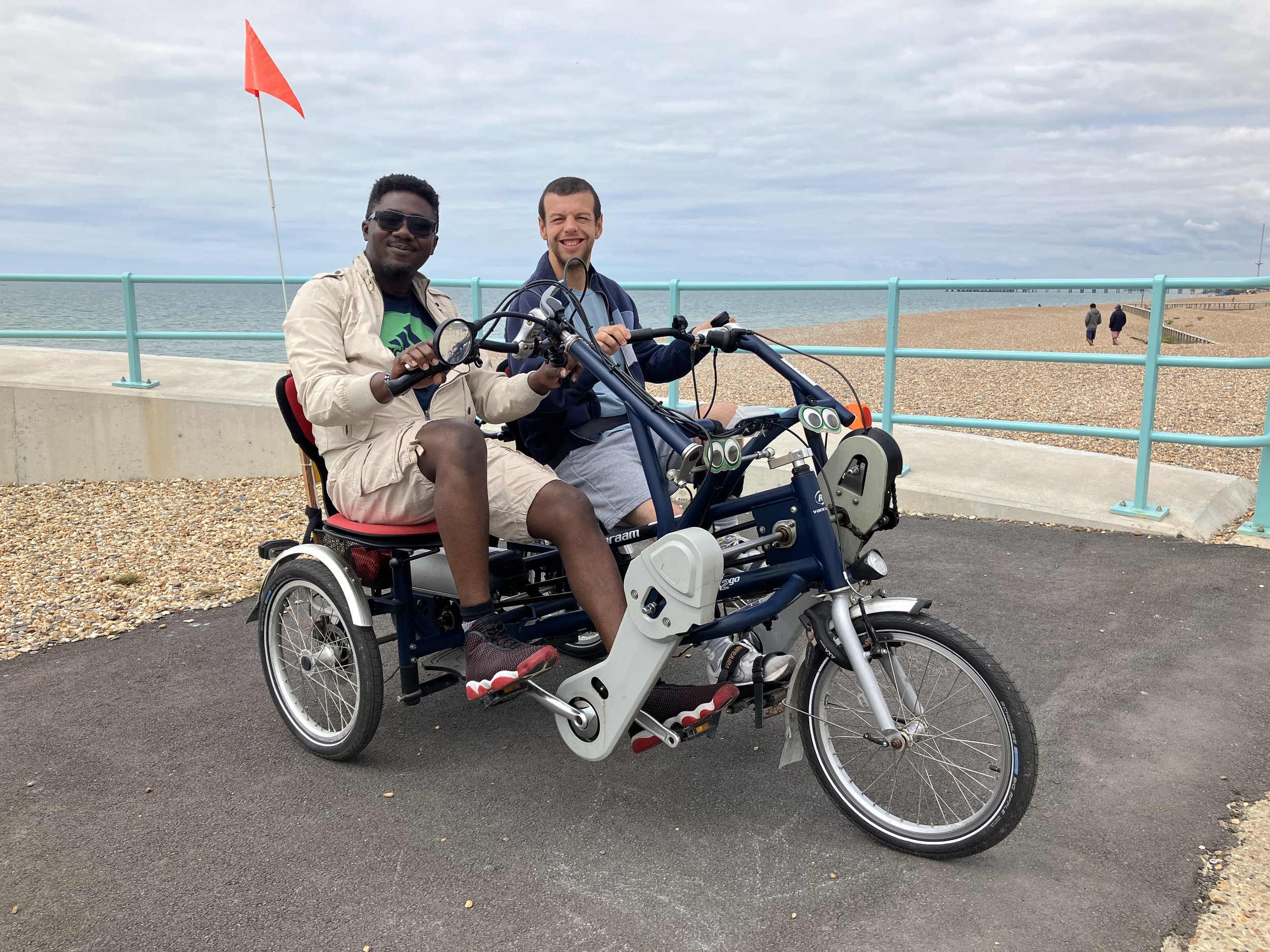 Side-by-side seated tandem cycle with 2 participants riding. Both smiling at camera. Pebble beach and seaside in the background.