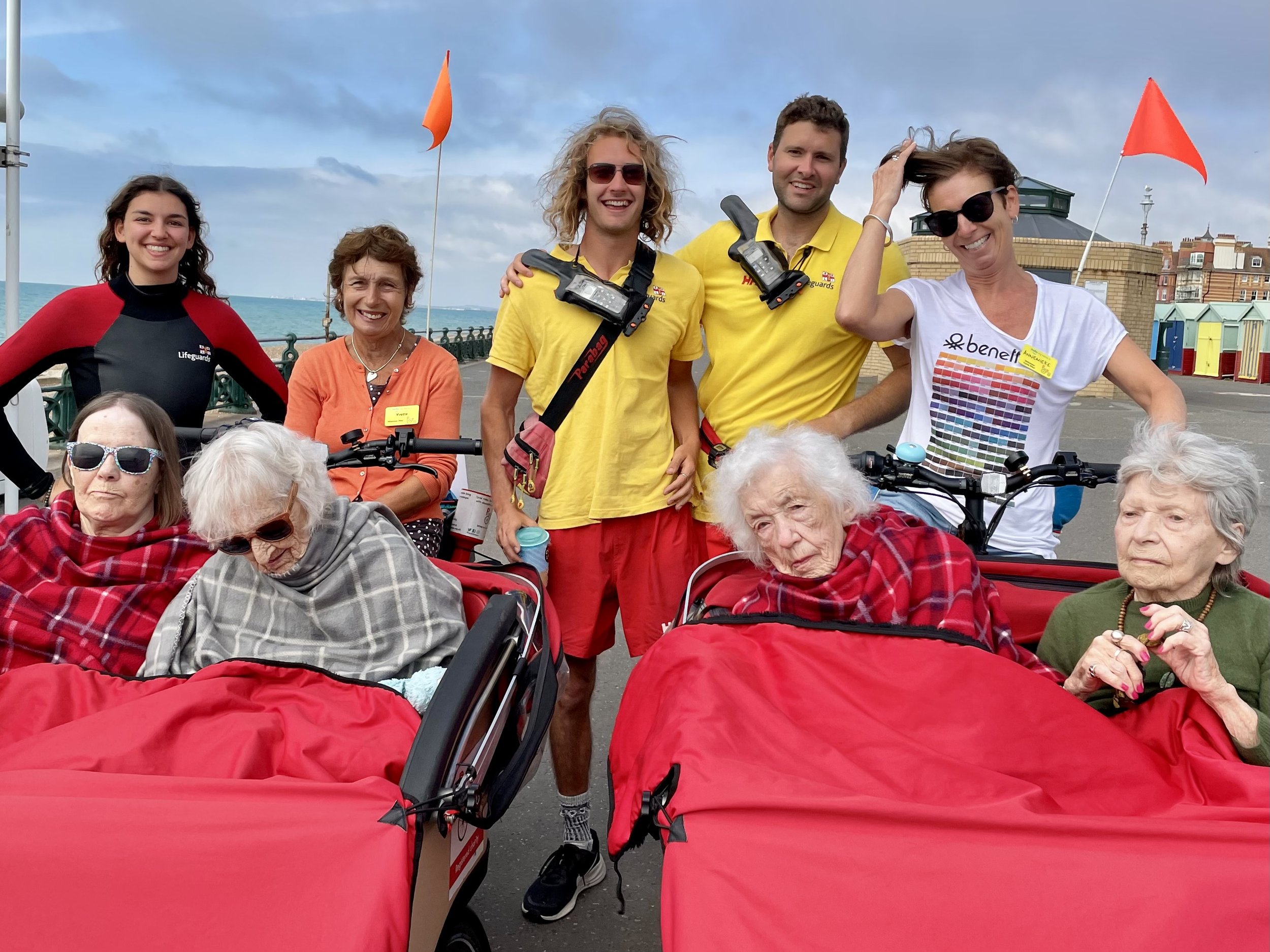 2 bright red trishaw cycles with 2 elderly participants seated in each. Our volunteer pilots stand behind them alongside three lifeguards, smiling at the camera. Seaside and beach huts in the background.