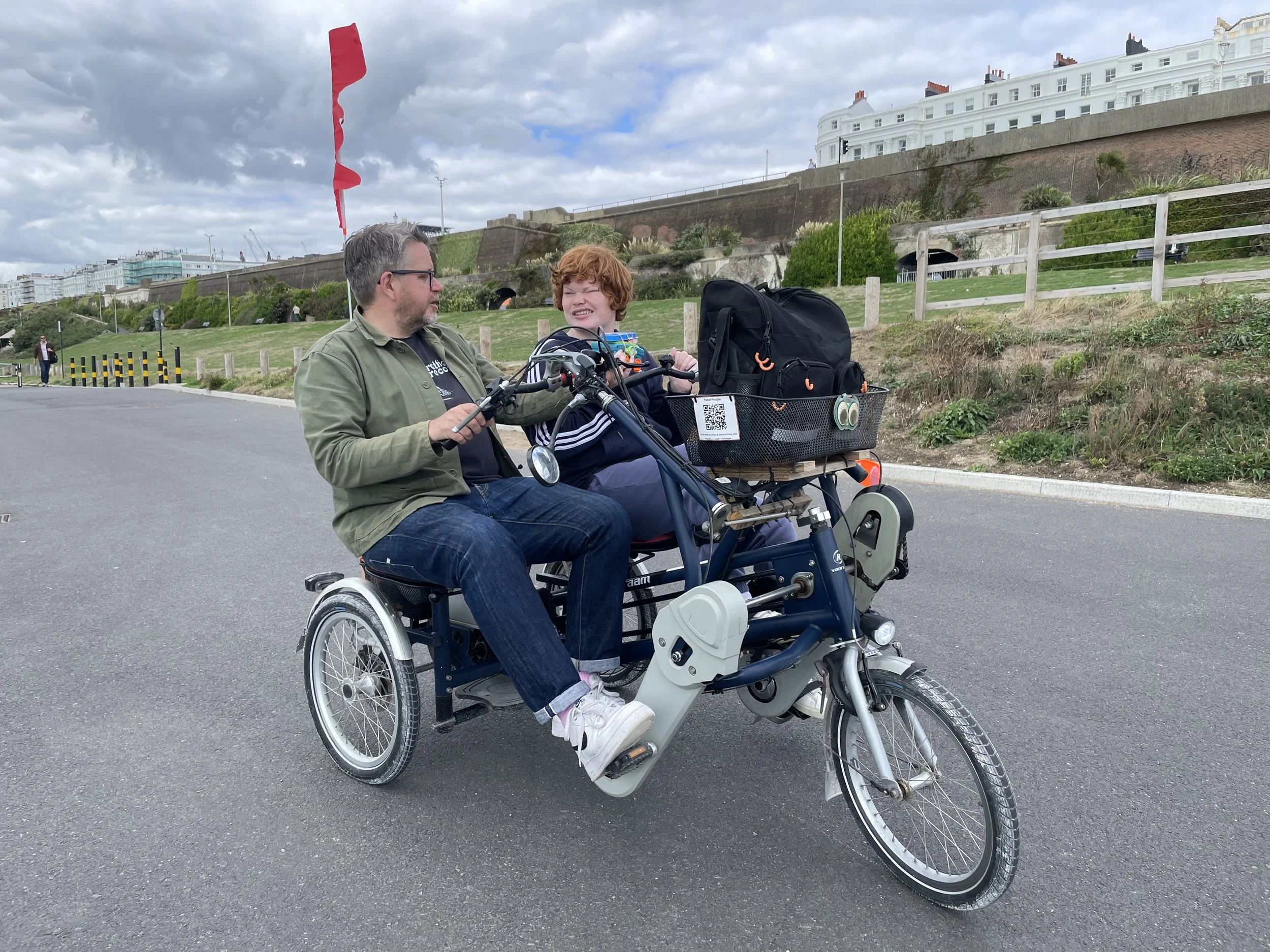 Two people look at each other as they sit on a side-by-side tandem. There is a grassy hill behind them and the sky is cloudy.