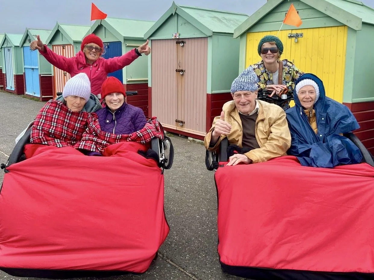 Two trishaws are parked in front of a row of brightly coloured beach huts. Each trishaw has two participants sitting on the front, warmly wrapped up in coats and blankets. The Cycle-Pilots sit behind them, one has their arms in the air. Everyone is s