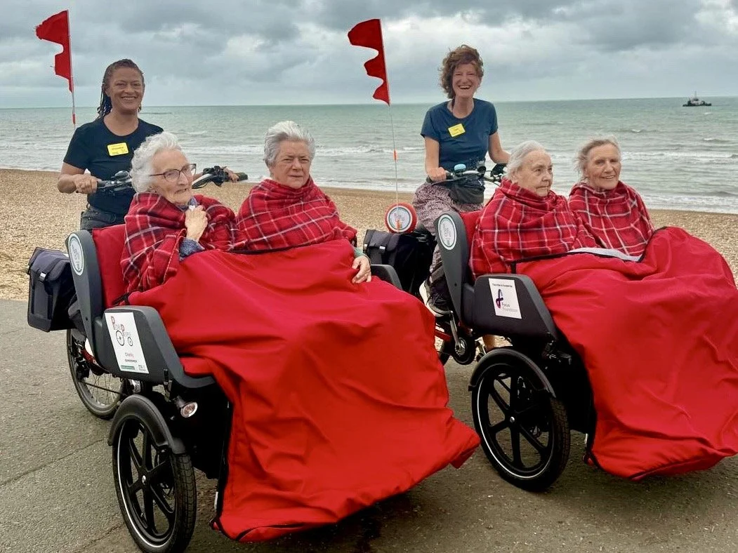 Two trikes are lined up next to each other, both with two people sitting in the front carriage and the riders behind them. They are wrapped up warmly, cycling along the seaside on a cloudy day.