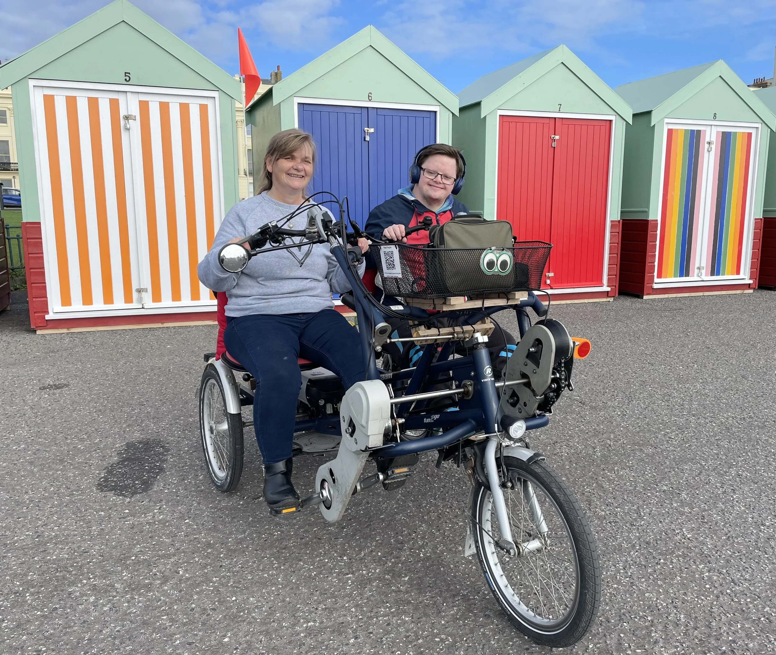 Two people riding a side-by-side tandem on a seafront promenade. They are smiling a the camera. Behind them are colourful and stripy beach huts. 