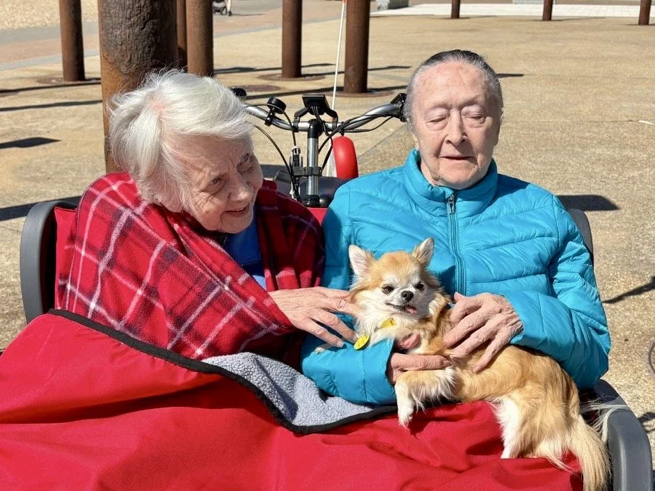 Two women sit in the front carriage of a trike. They are stroking a small chihuahua dog.